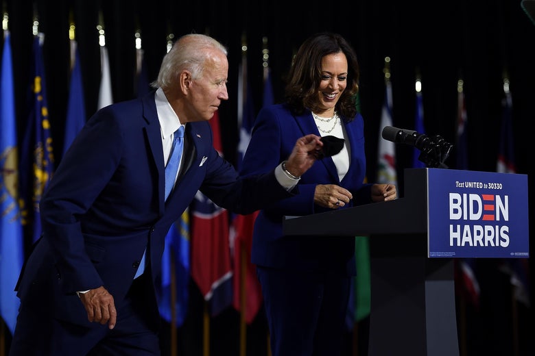 Joe Biden, holding a black face mask, leans toward a smiling Kamala Harris, who is standing behind a lectern and microphone, a row of flags behind them.