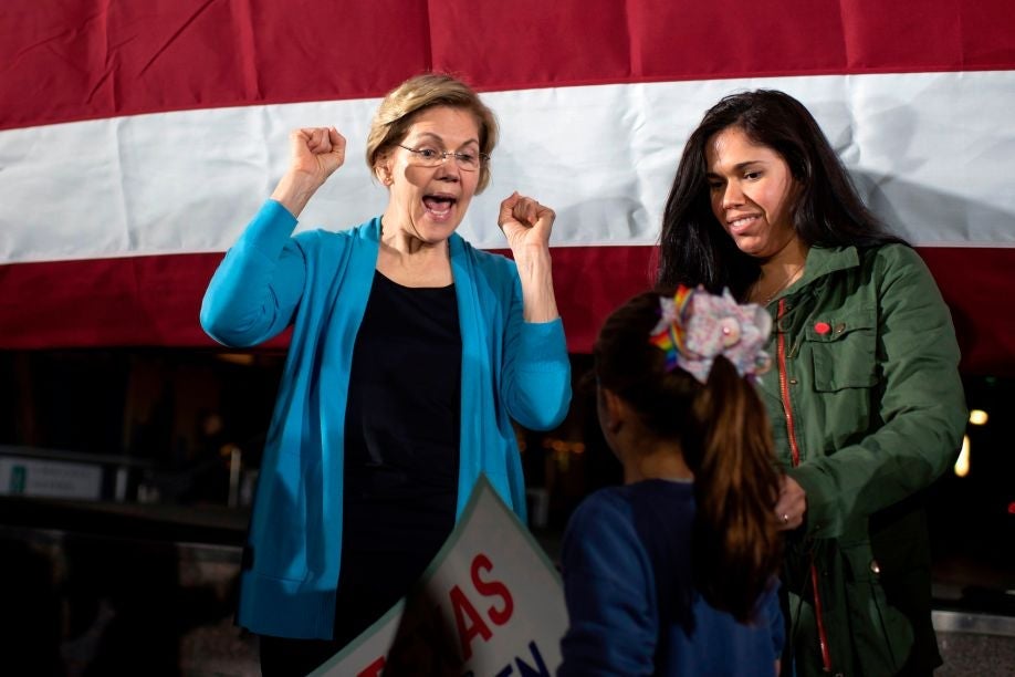 Warren makes a "hooray" gesture while speaking to a young girl, against the backdrop of a large American flag.