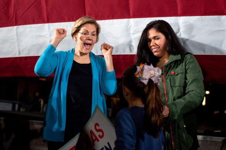 Warren makes a "hooray" gesture while speaking to a young girl, against the backdrop of a large American flag.