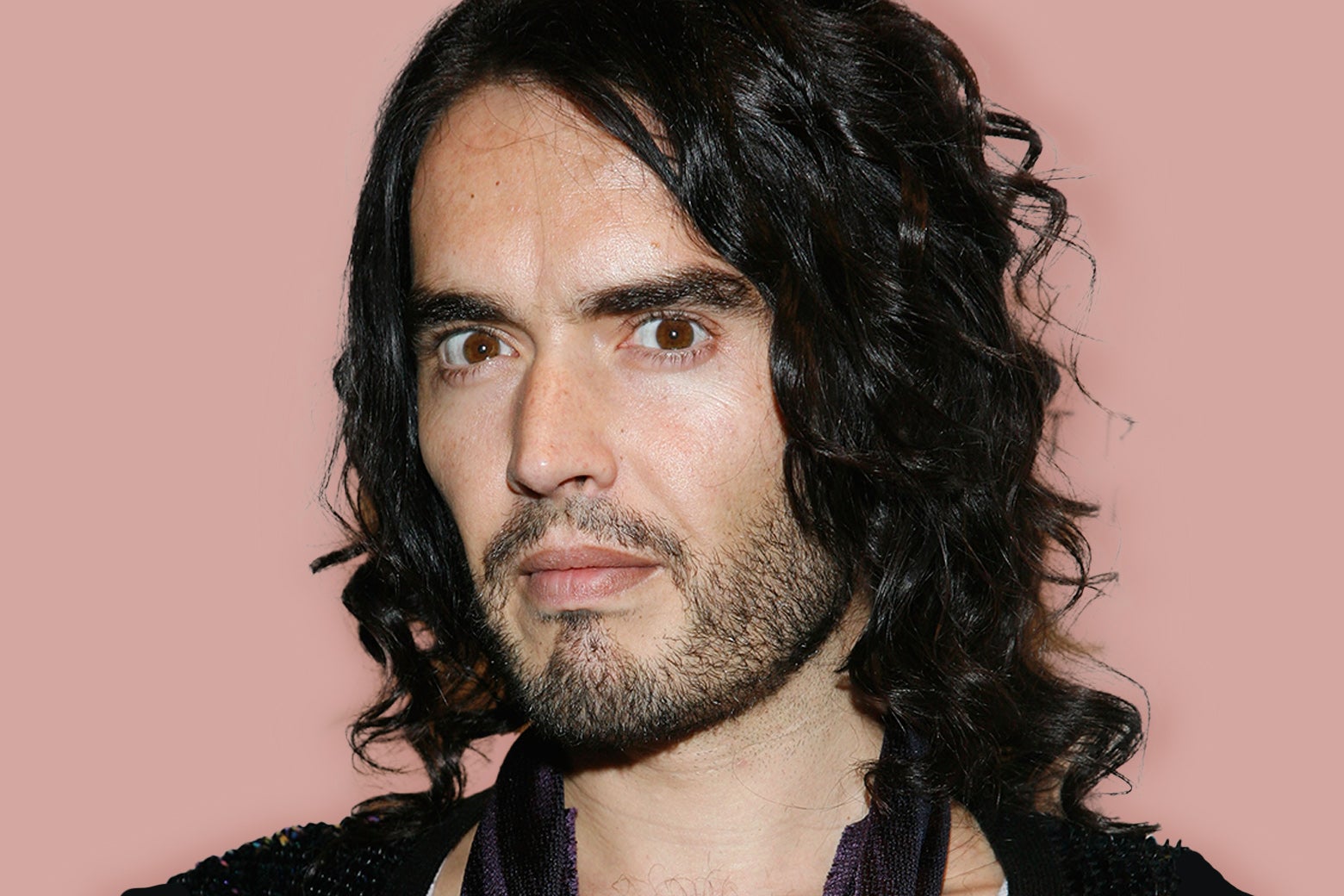 Russell Brand looks into the camera over a pink background.