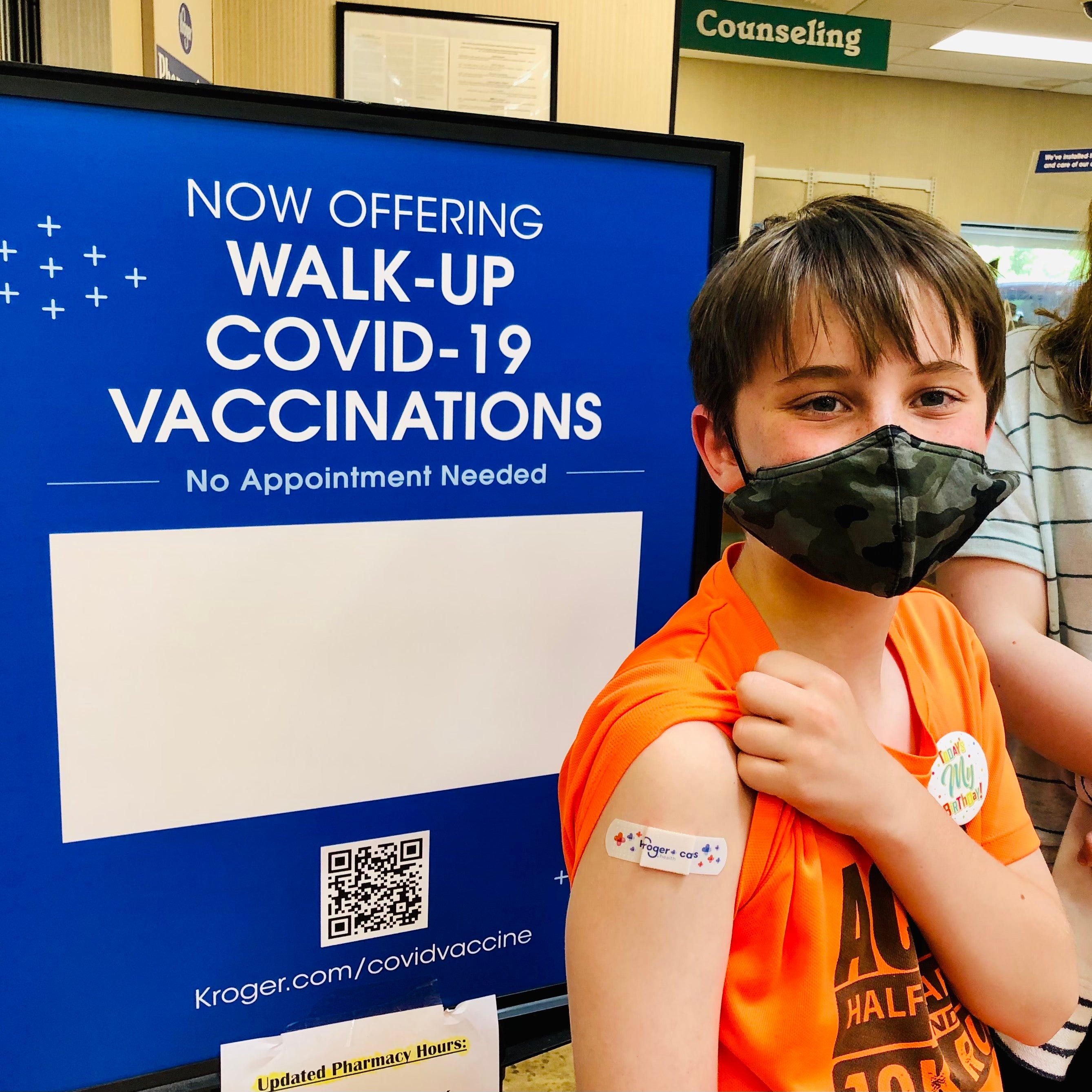 A masked boy in a pharmacy lifts his sleeve to show a band-aid. A masked girl stands behind him.