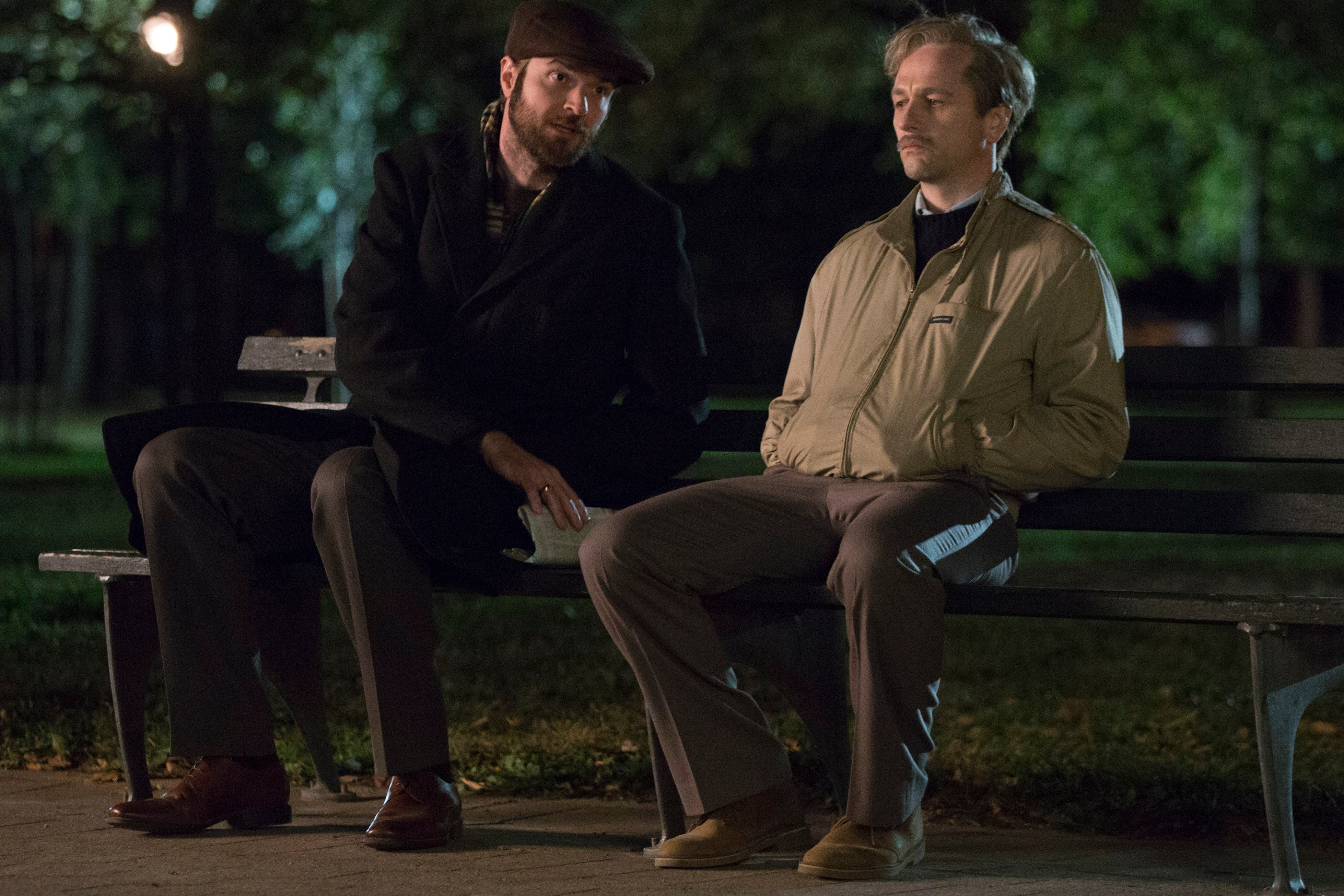 Philip (Matthew Rhys) and Oleg (Costa Ronin) have a conversation on a park bench in FX's The Americans. They are both young men. Oleg, to the left, has a short beard and wears dark clothes and a flat cap. Oleg, to the right, wears a khaki jacket, pants, and shoes.