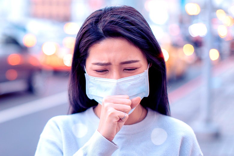 A woman in a surgical mask coughs into her fist.