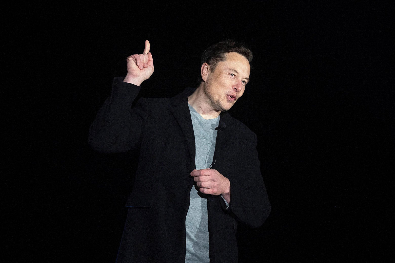 Elon Musk points up while speaking from a stage with a black background.
