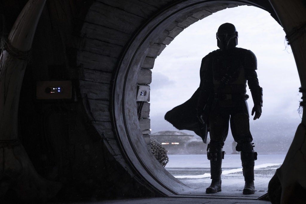 A silhouette of a person wearing armor, a helmet, and a cape standing in a round doorway.