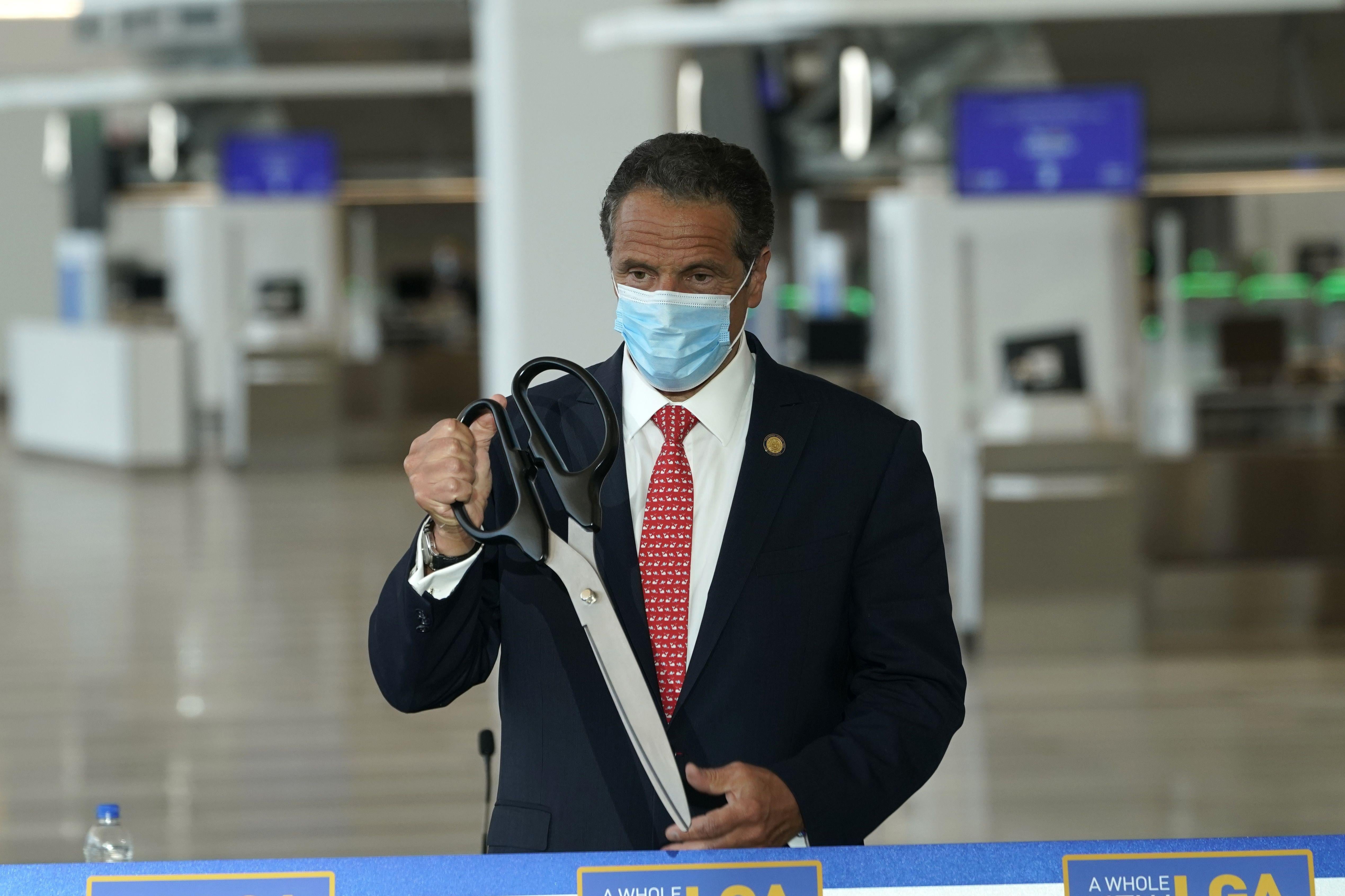 Cuomo wearing a surgical mask and holding a giant pair of scissors as he stands next to a ribbon in the terminal.