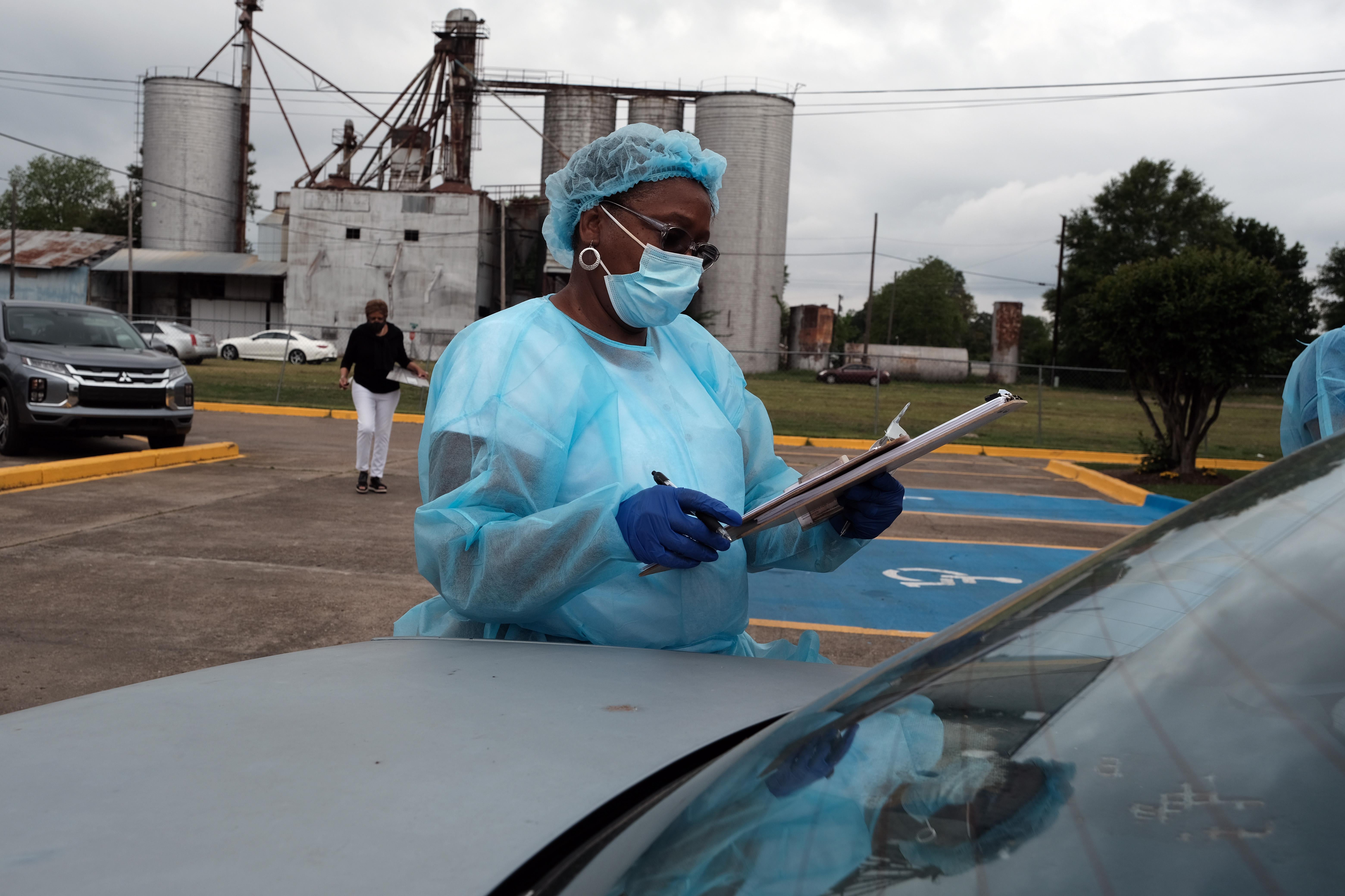 A woman in a surgical gown and a mask looks at a clipboard while standing near a car.