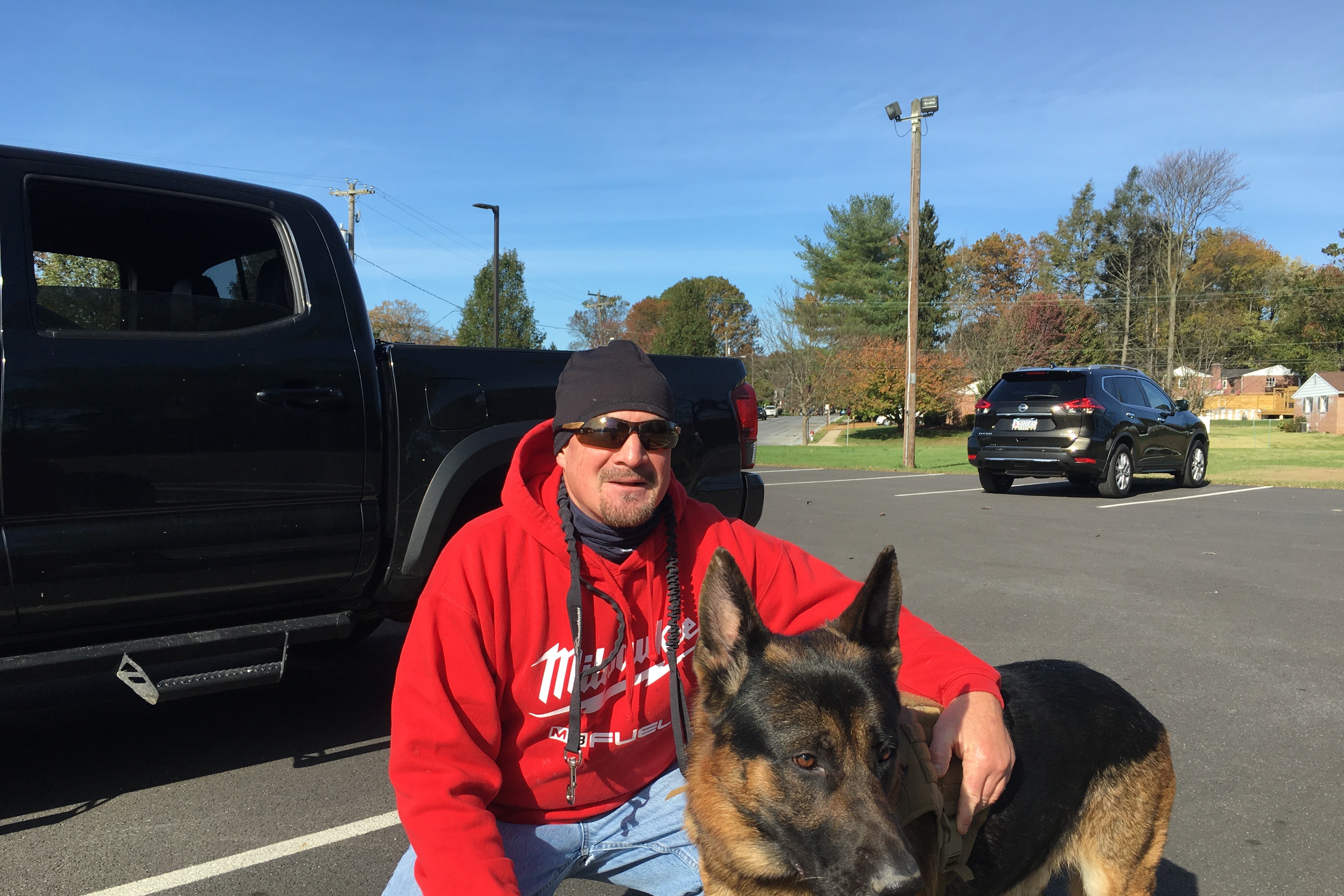 A man wearing a red hoodie crouches beside his dog in front of a pickup truck in a parking lot