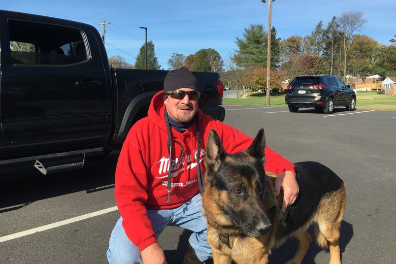 A man wearing a red hoodie crouches beside his dog in front of a pickup truck in a parking lot