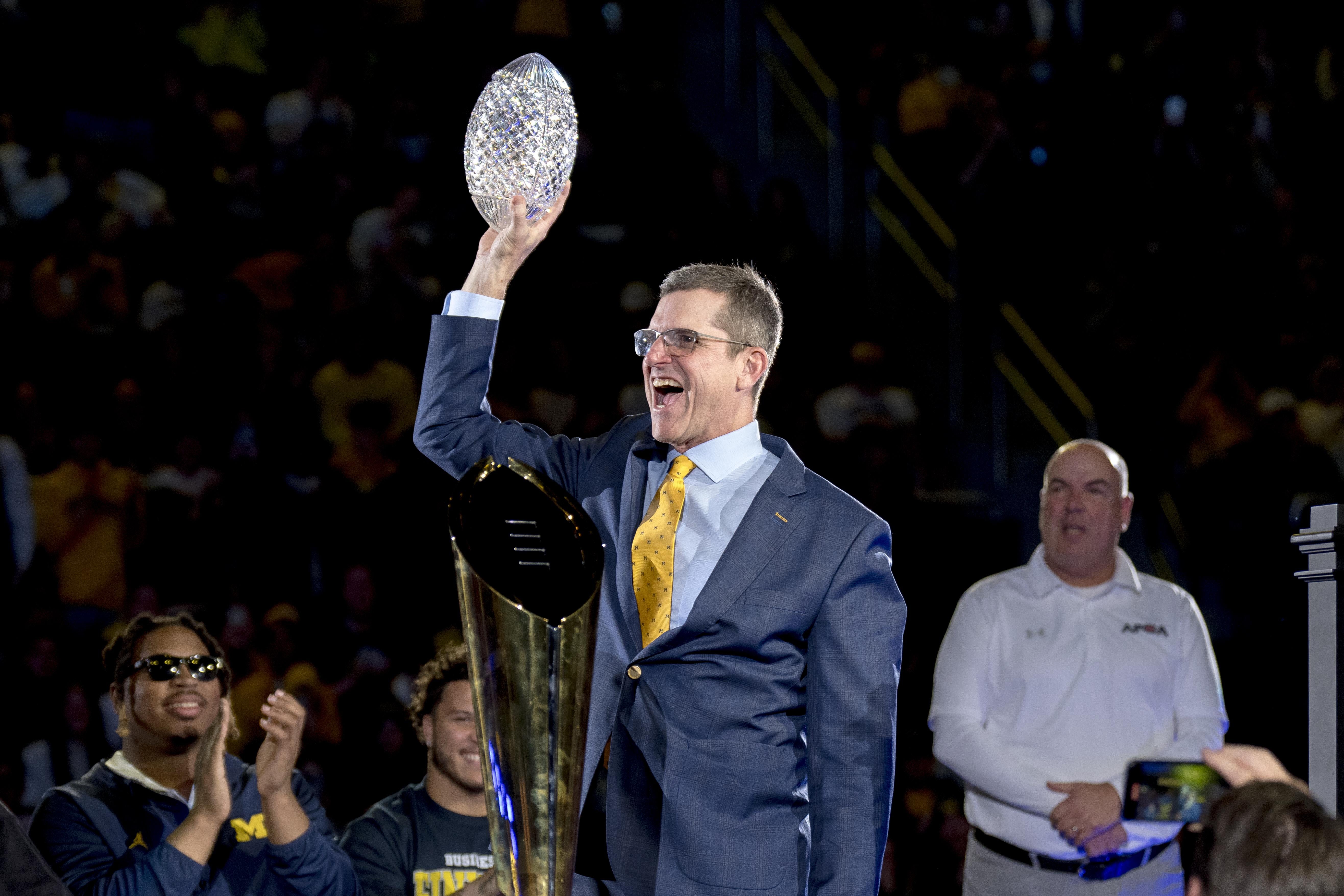 Harbaugh laughs onstage, holding a glass/crystal football in the air with his right hand, and a large trophy standing in front of him.