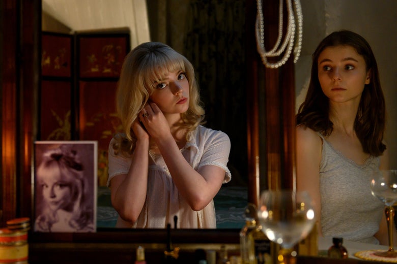 Anya Taylor-Joy stands in front of a mirror in long, blond, 1960s hair, removing an earring. Thomasin McKenzie, in a gray tank and brown hair, looks on. Is she in the mirror?