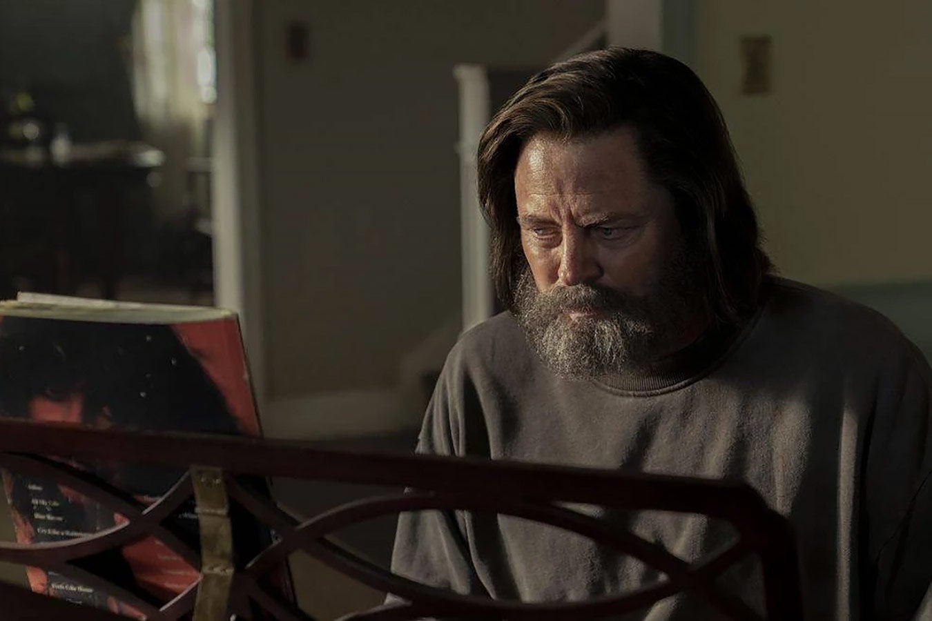 A white man with an overgrown beard sits at a piano in a dim room.