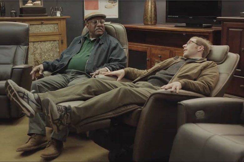 Two men sit on leather chairs: One is wearing a gray hat and green shirt, and the other has a brown jacket. The one in the brown jacket is reclining.