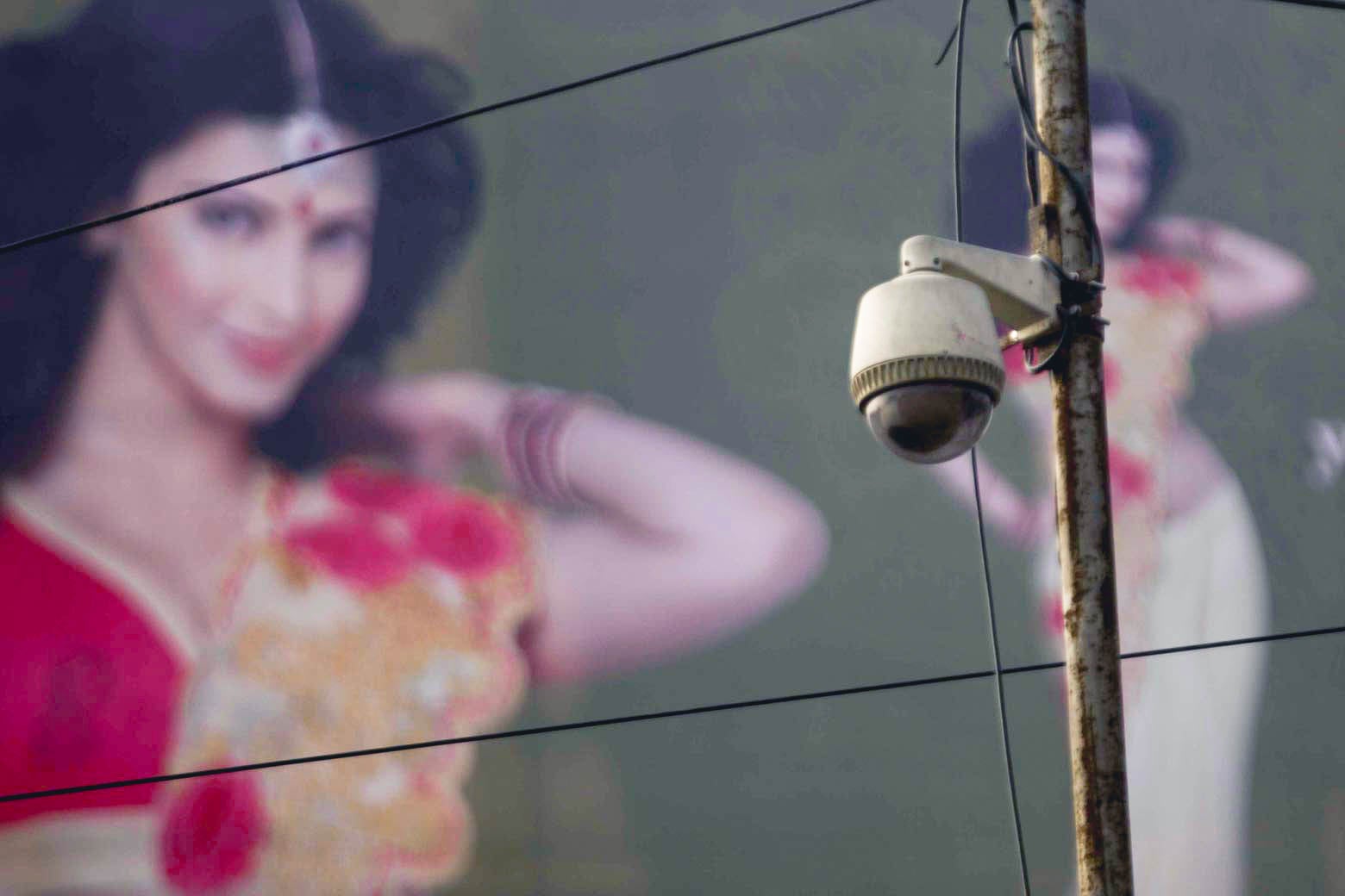 A CCTV camera on a pole in front of a mural of a woman in traditional Indian dress.
