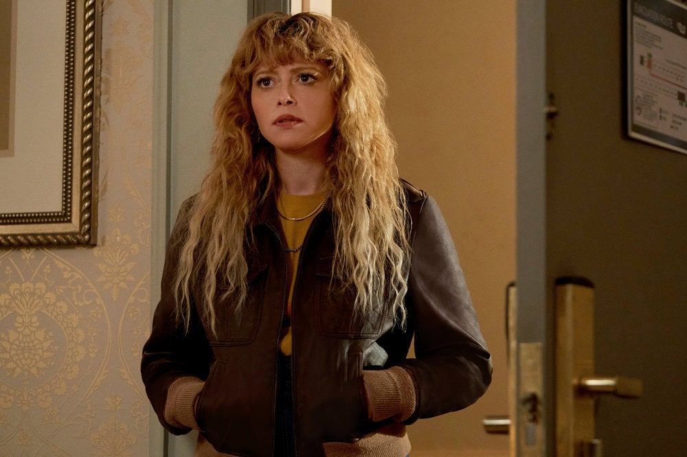 She stands in a doorway in a stylish brown bomber jacket and gold necklaces, a concerned look on her face, her blonde curls raining down around it
