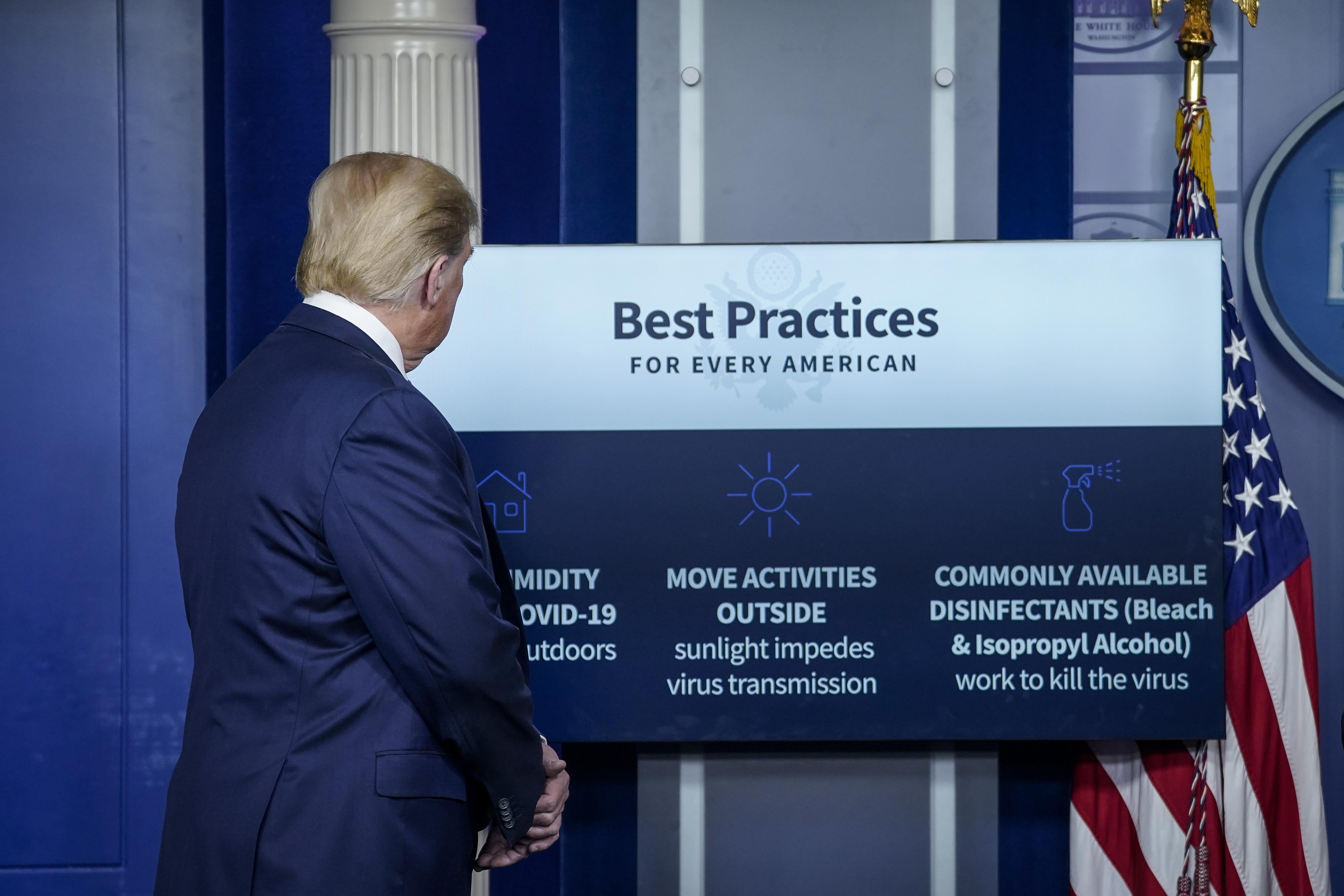 Trump looks at a poster listing "Best Practices for Every American," including "Commonly Available Disinfectants"