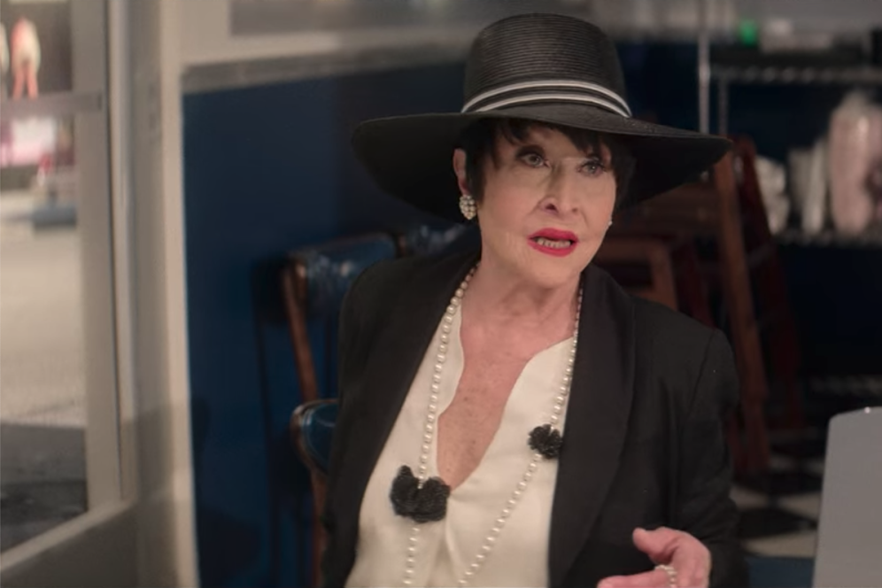 Chita Rivera sits at a diner table, singing while drinking a martini.