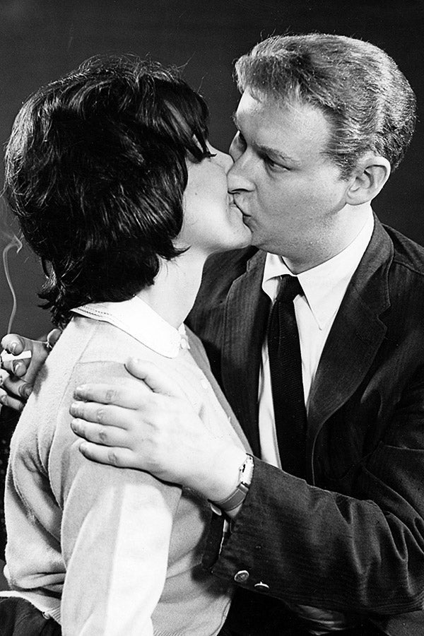 Mike Nichols planting a big kiss on Elaine May while holding a cigarette in his right hand.