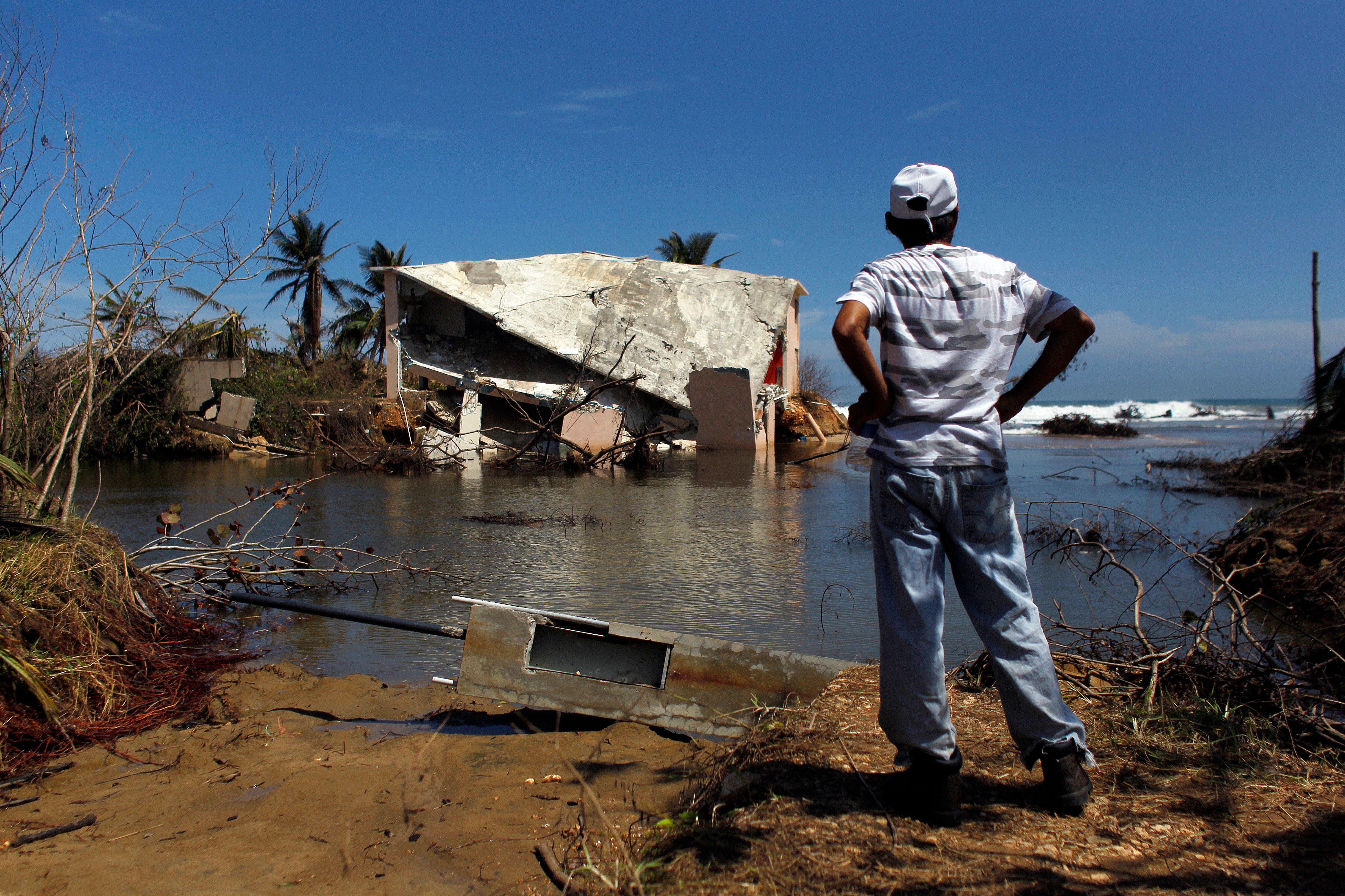 A man with his hands on his hips stands looking at a damaged house surrounded by water.