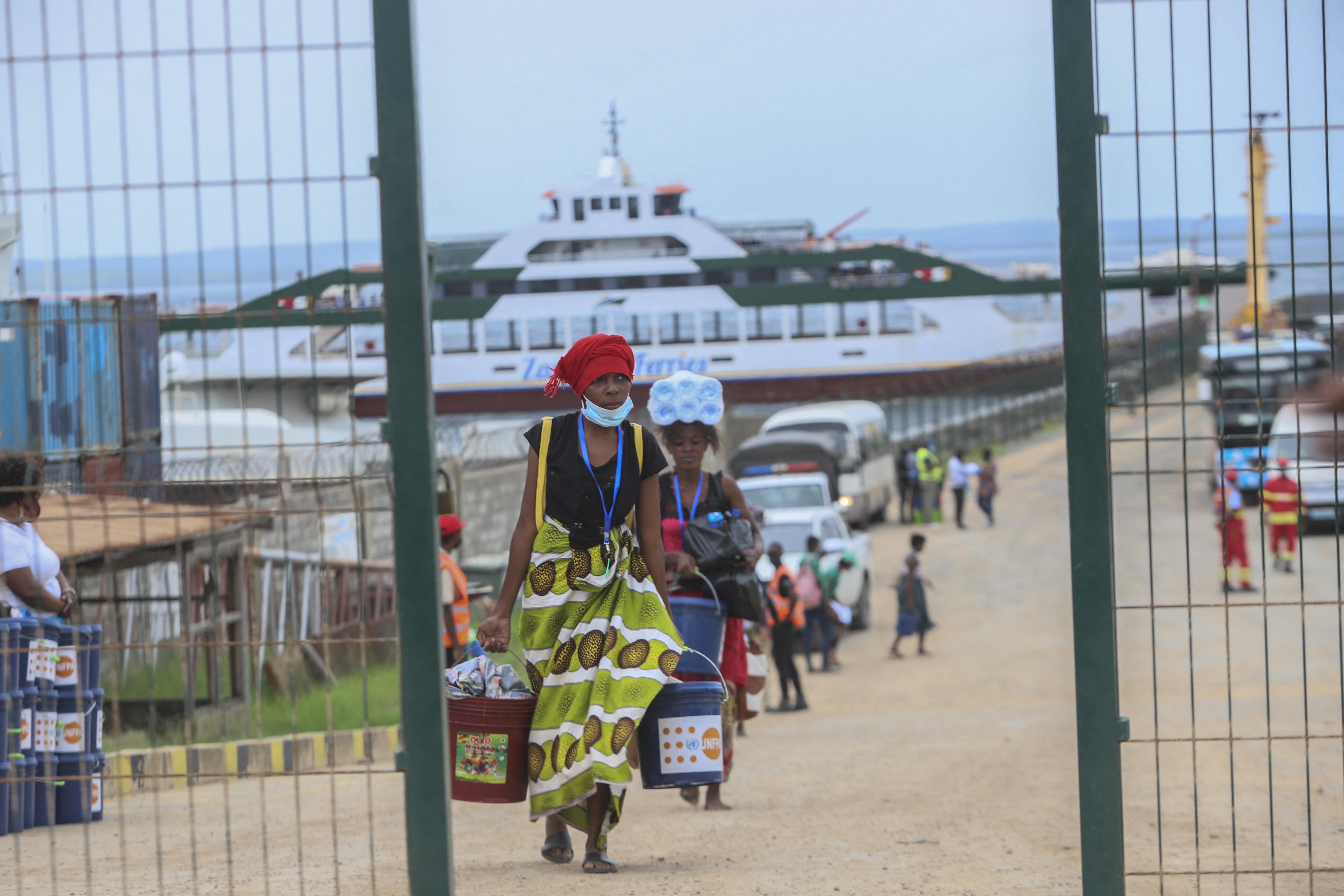 With a large ship on the water in the background, people carrying possessions walk up a dirt path toward an open gate in a fence.  