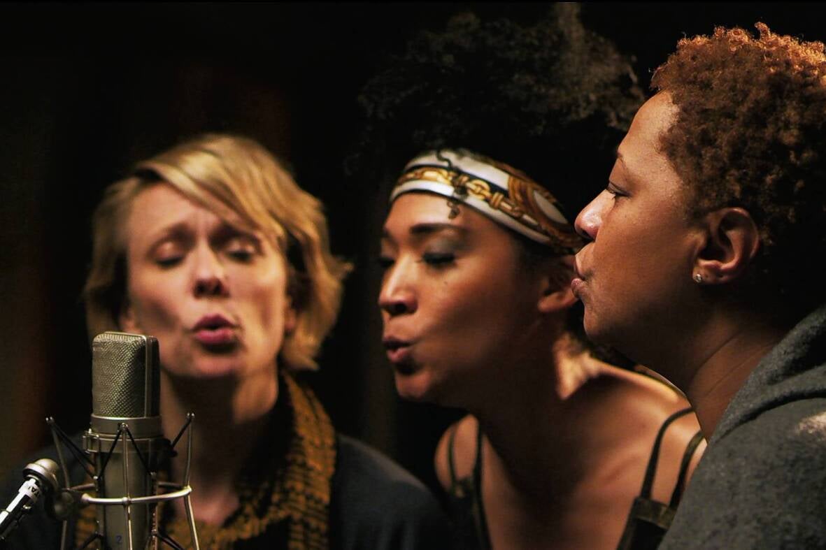 Jo Lawry, Judith Hill, and Lisa Fischer sing with rounding mouths into a single microphone in a recording studio.