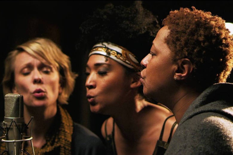 Jo Lawry, Judith Hill, and Lisa Fischer sing with rounding mouths into a single microphone in a recording studio.
