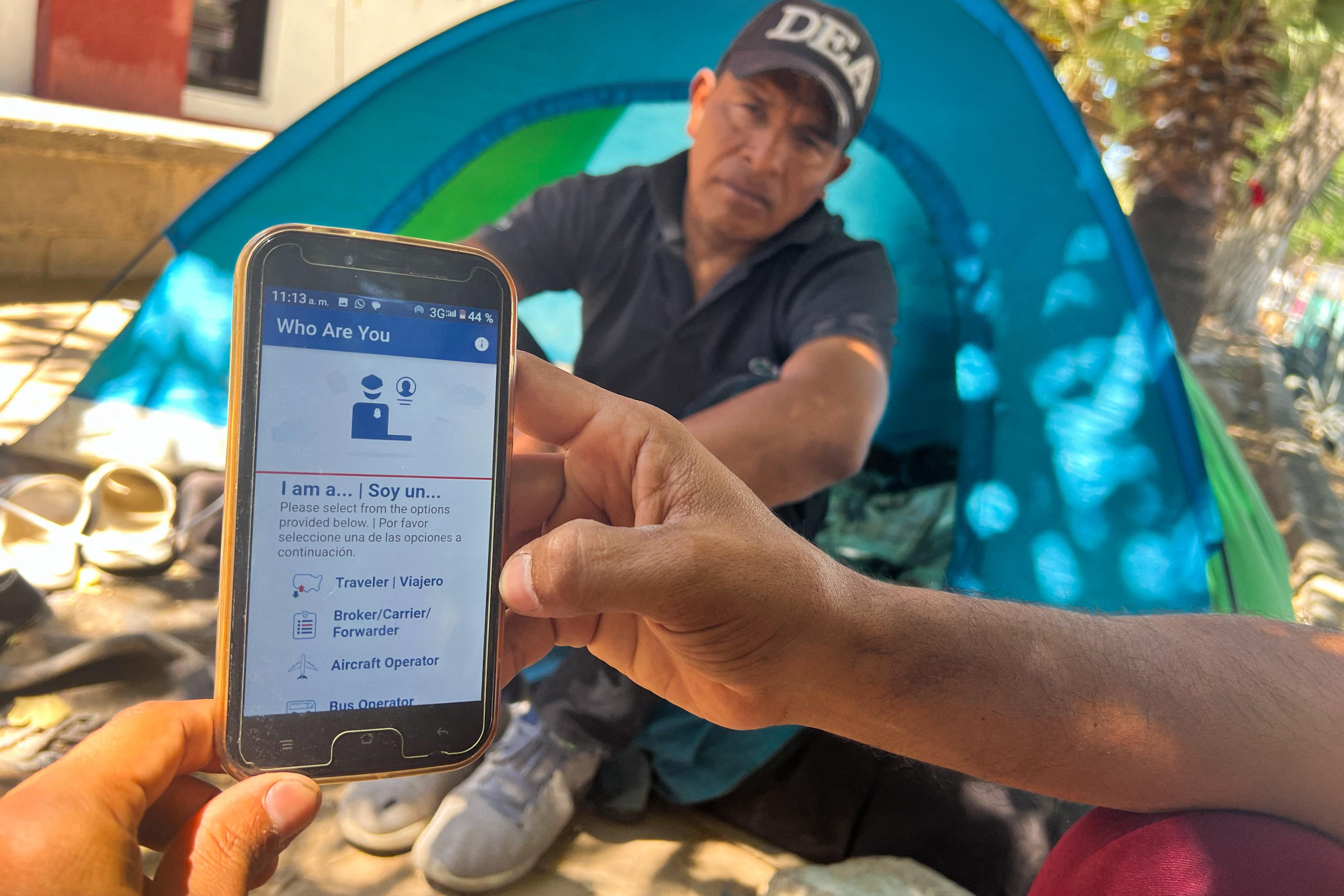 A DEA agent sitting in front of a tent stares skeptically at a person. All you can see of that person is one arm and a second hand holding up a phone with an app reading "WHO ARE YOU."