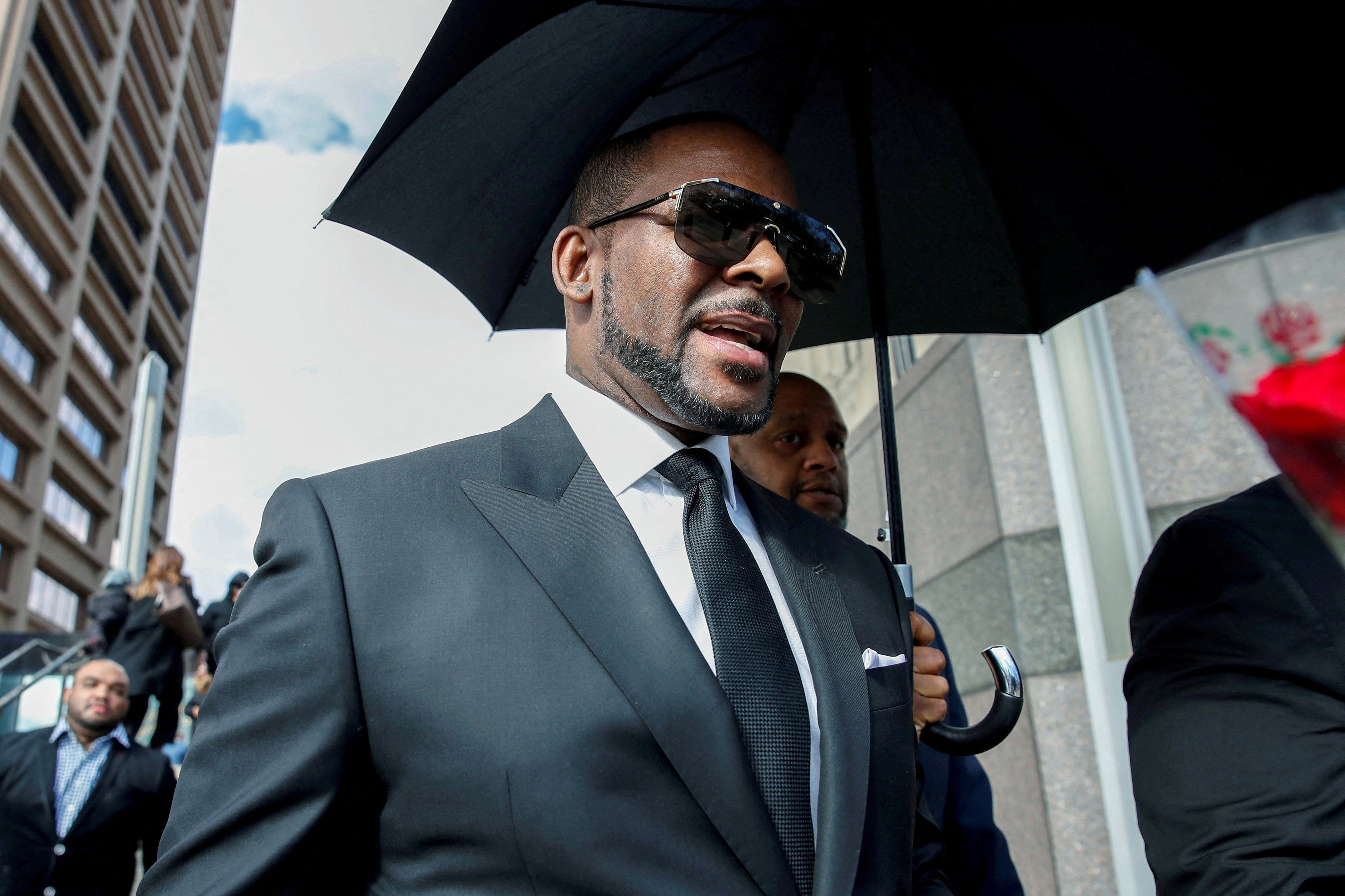 R. Kelly wearing a suit and sunglasses and walking under an umbrella