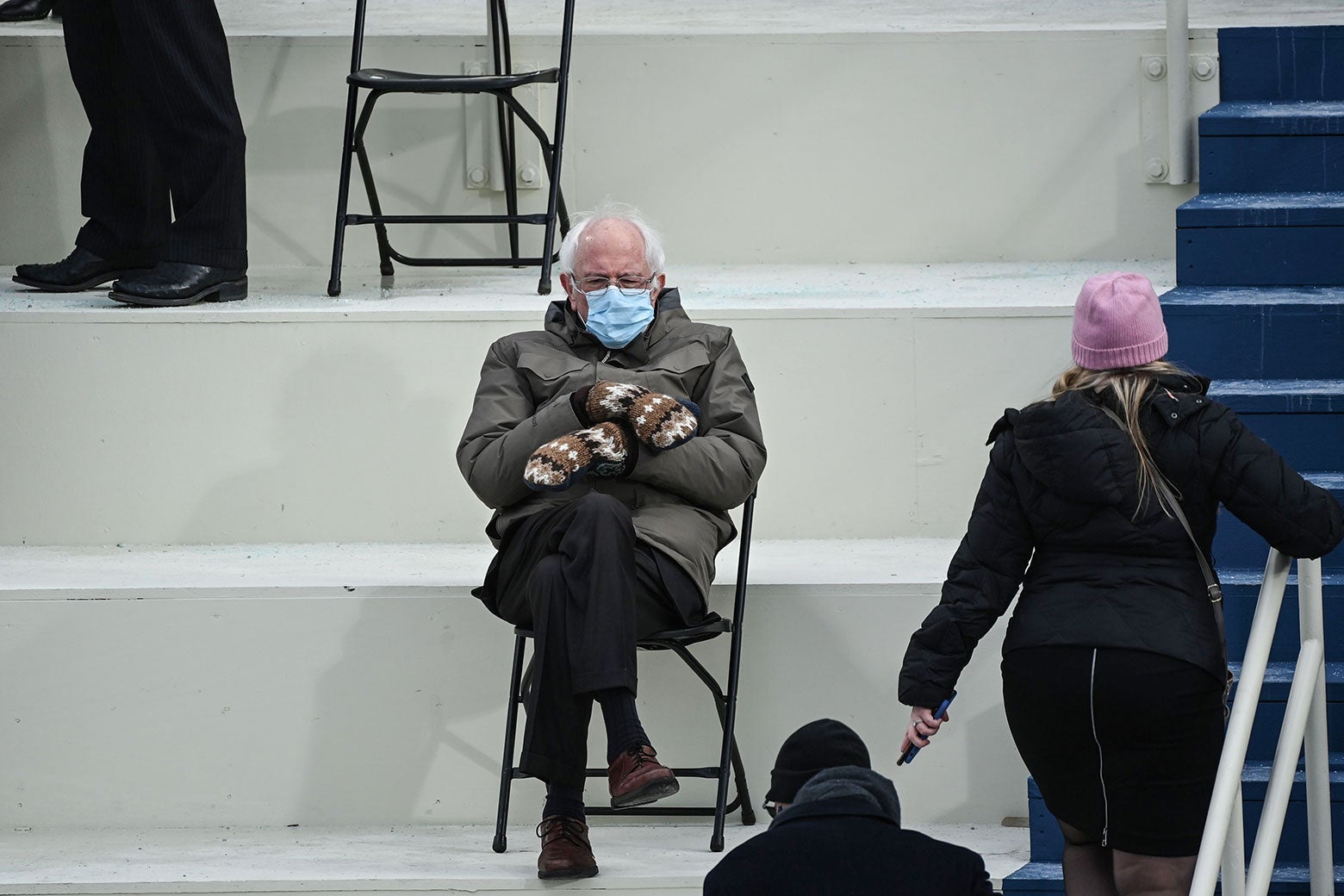 Bernie Sanders sits cross-legged in a folding chair in an olive-green coat and knit mittens.