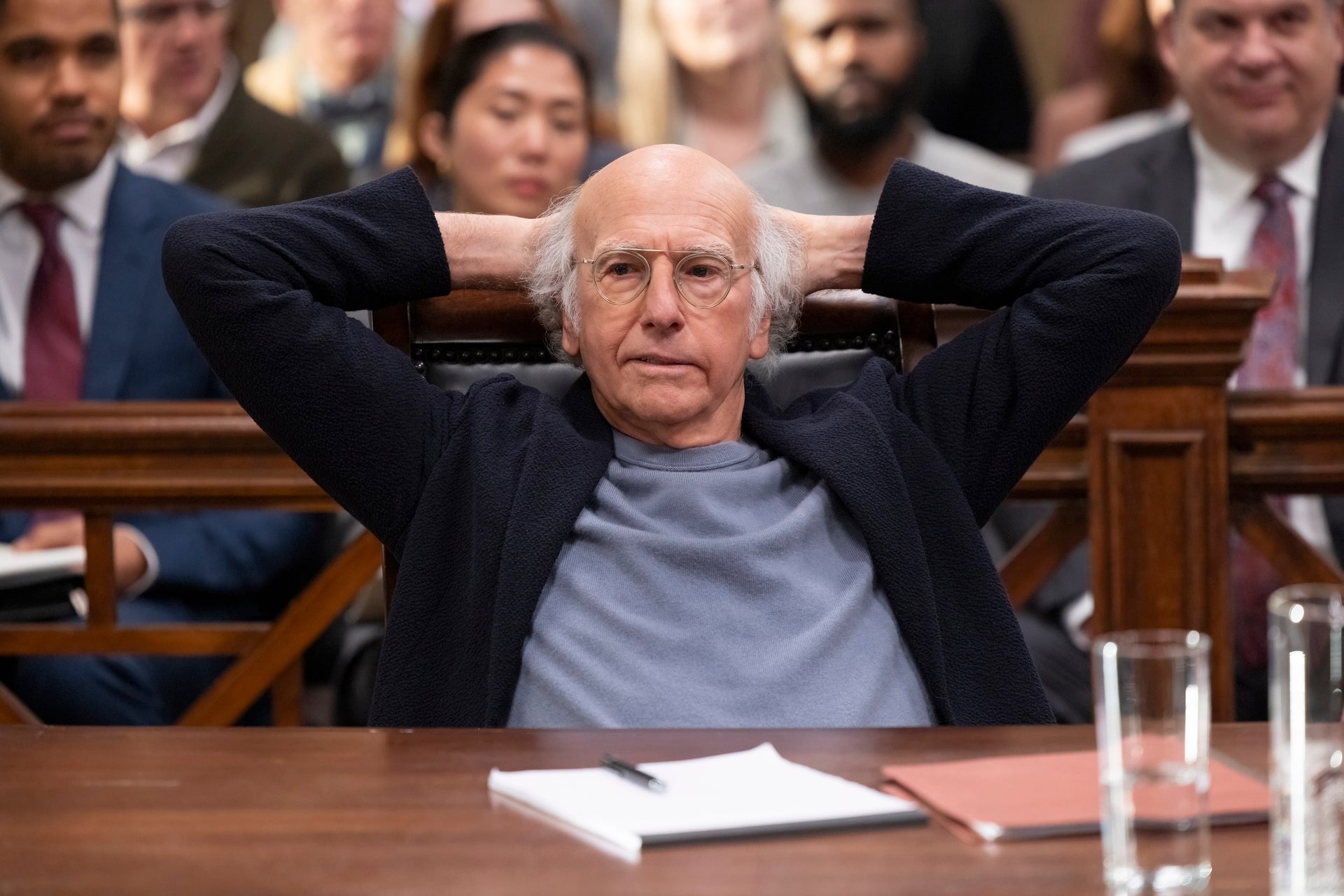 A still shows Larry David leaning back behind the defendant's table, his hands folded behind his head, looking defiant.