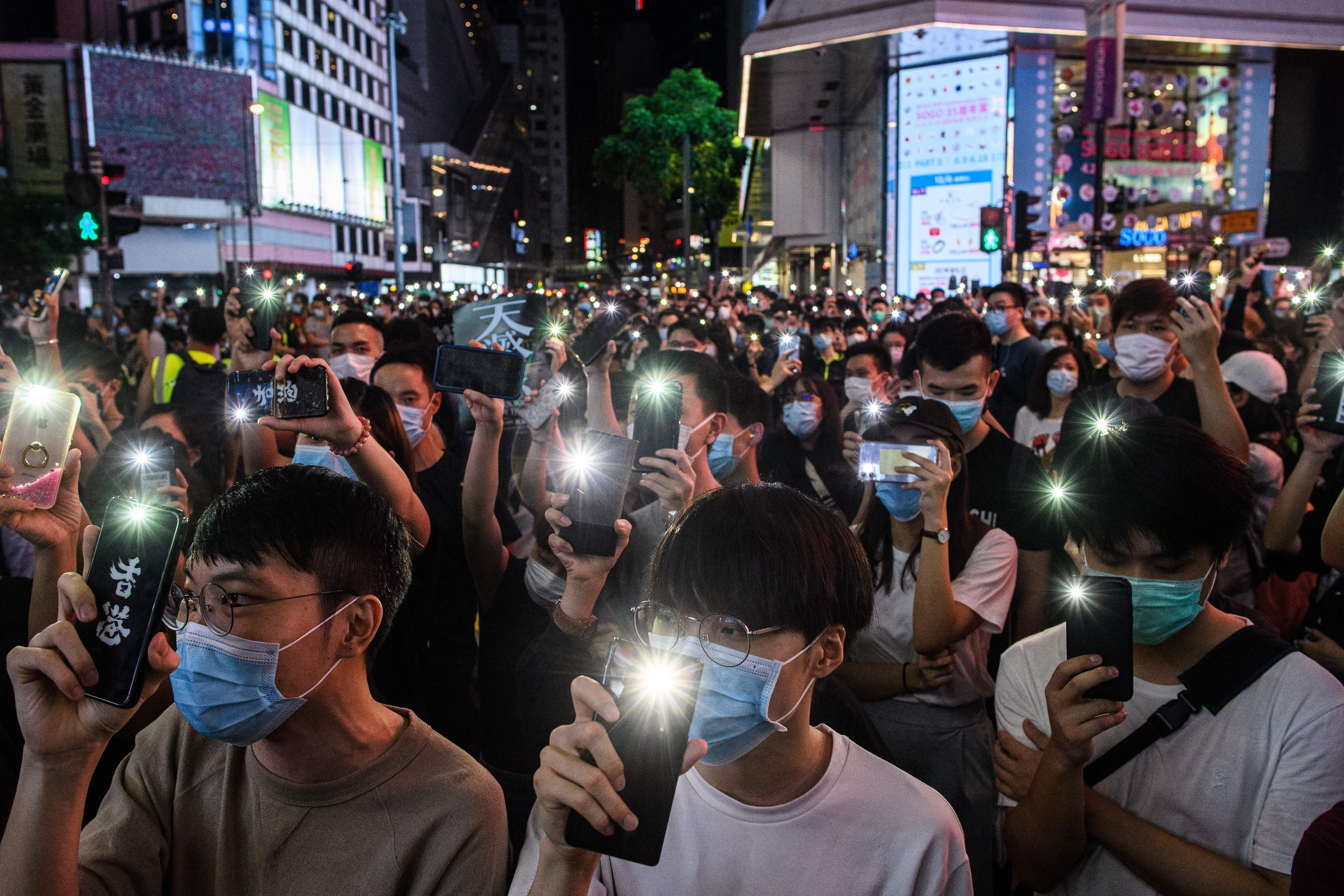 A large group of people stand in a city street at night, holding up phones with the flashlights turned on.
