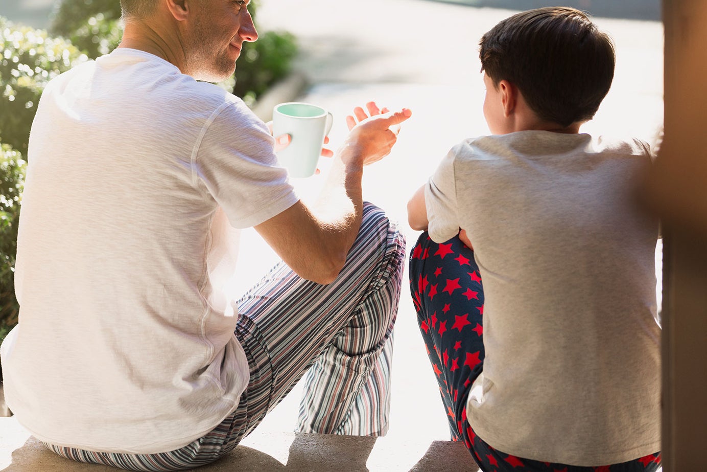 A man and boy sits on the front step of a house having a conversation while wearing pajamas.