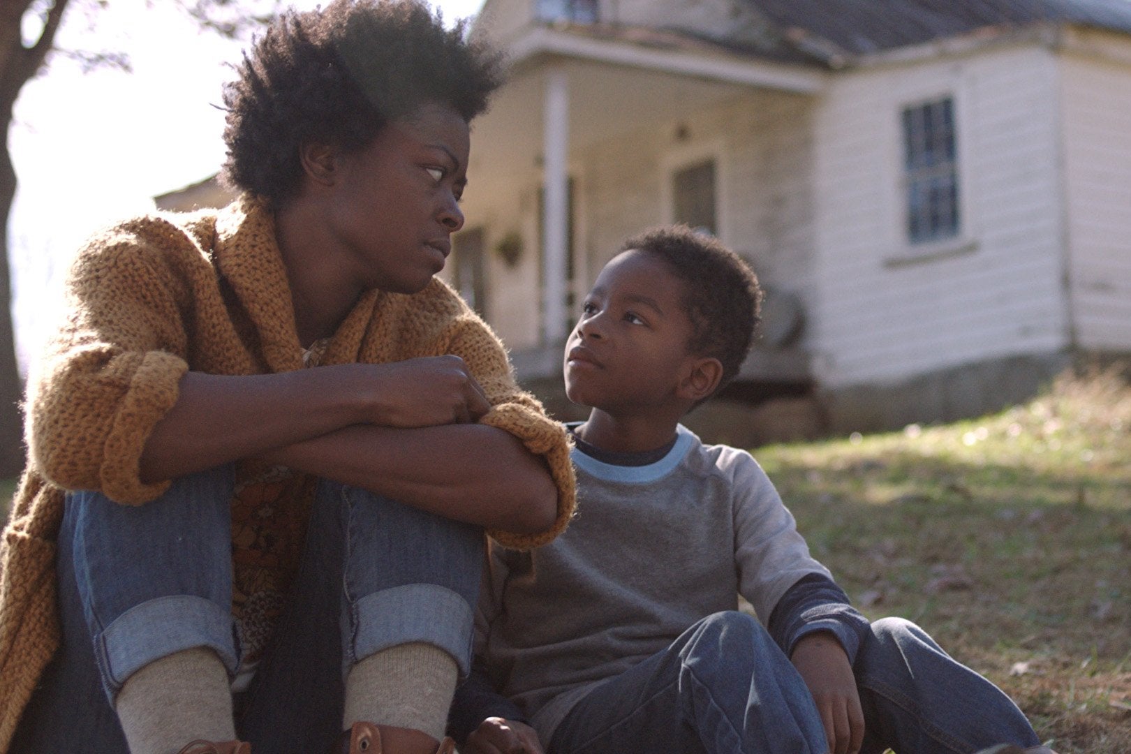 A black woman sits on a lawn with a young black boy in front of a slightly worn down house.