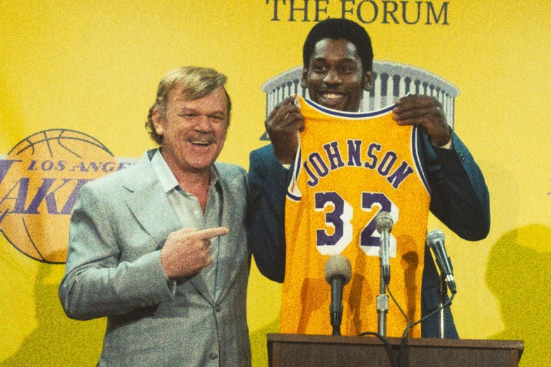 Reilly as Buss smiles and points at Isaiah as Johnson smiling and holding up his Lakers jersey at a podium in a grainy still from the show