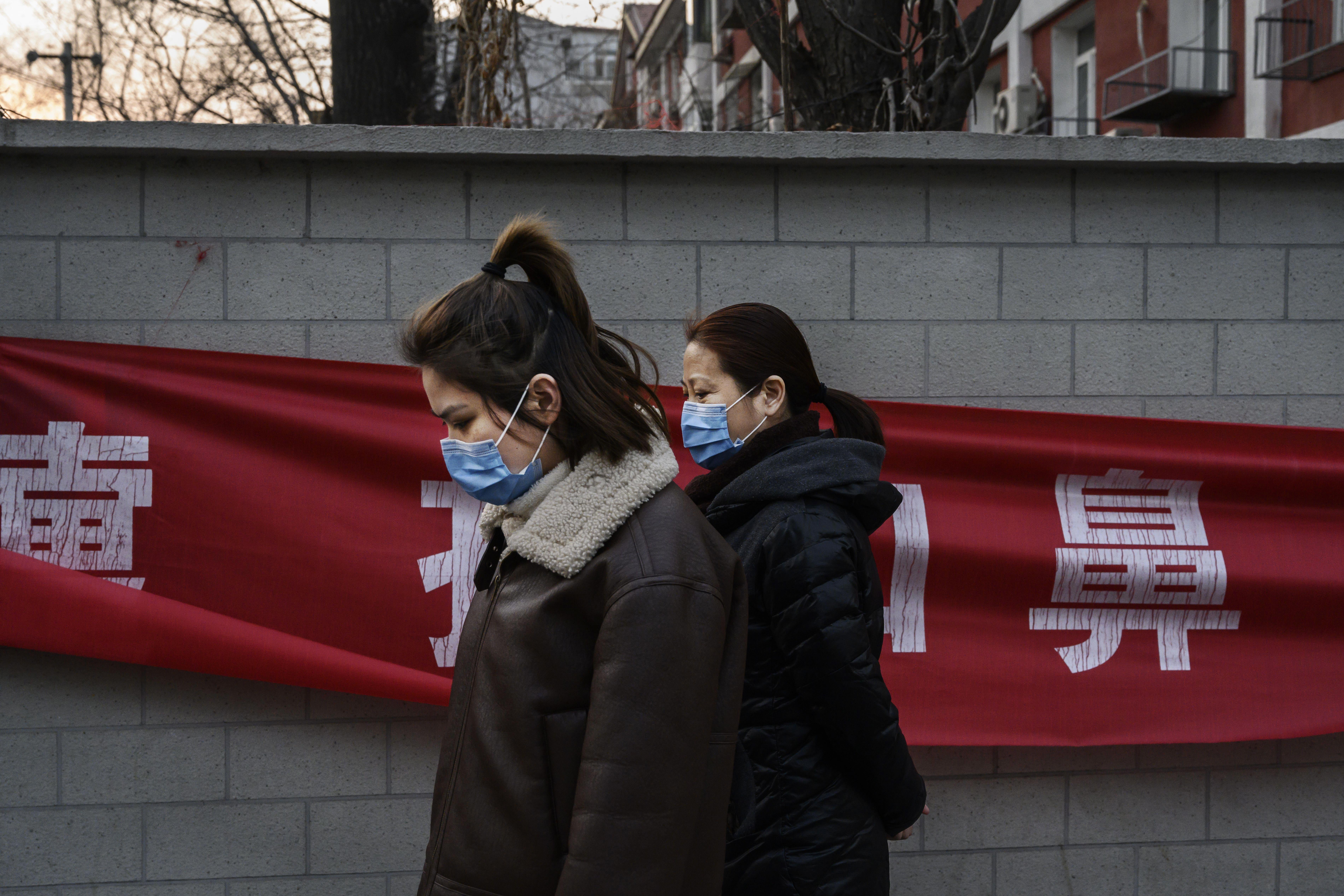 Two women wearing parkas and masks walk by a red banner.