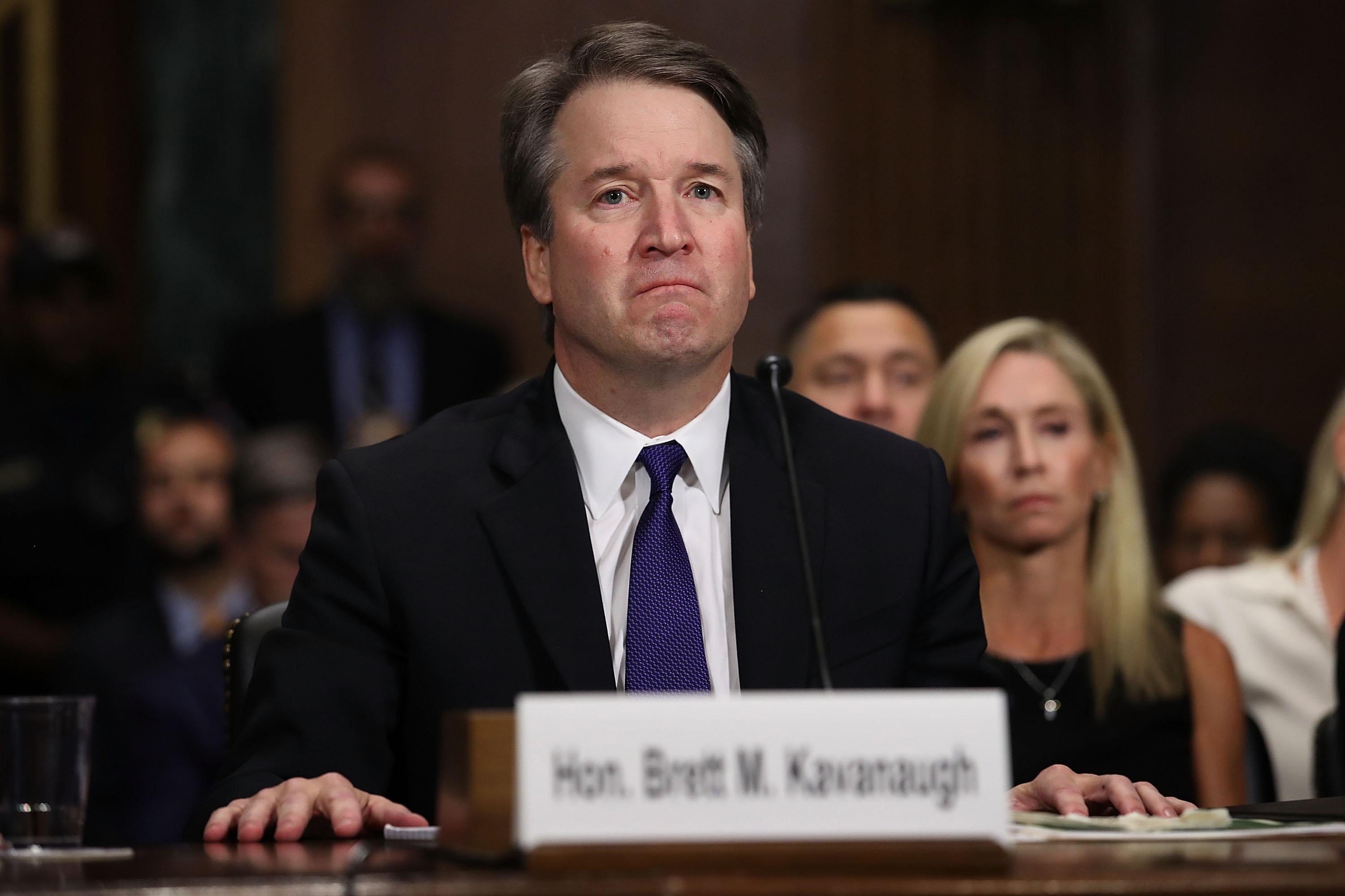 Kavanaugh, wearing a suit and seating at a table in front of an audience for the hearing, looks unhappy.