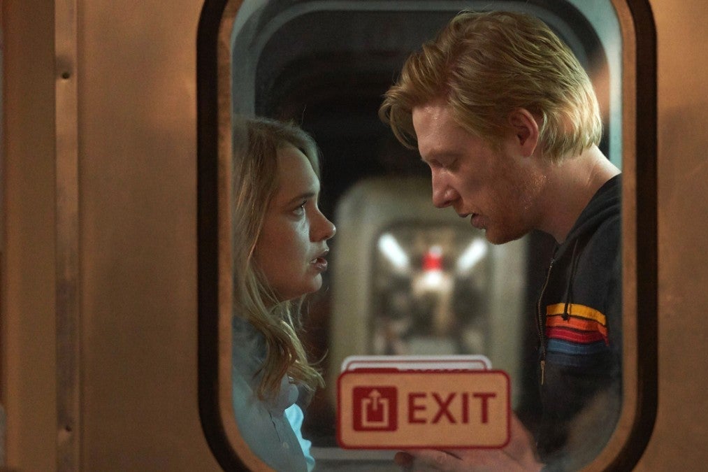 Merrit Wever and Domhnall Gleeson, sharing an intimate moment, seen through the gritty window of a train that has an Exit sign on it
