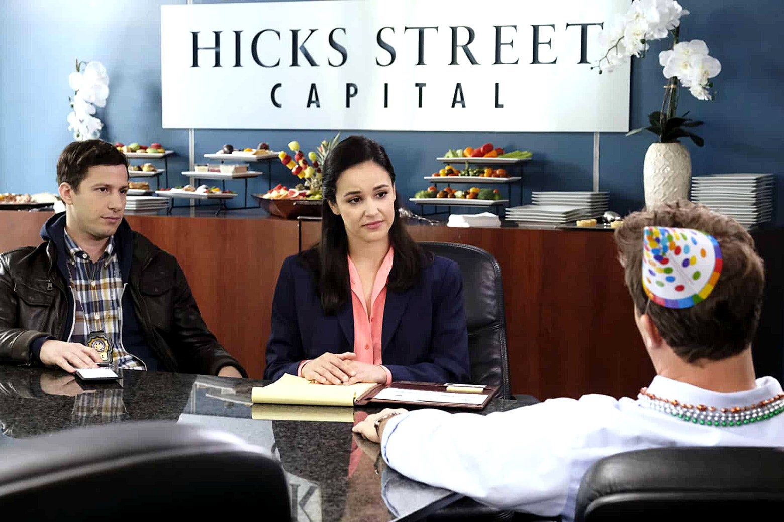 Andy Samberg and Melissa Fumero as Jake and Amy in a scene from Brooklyn Nine-Nine. Jake and Amy are seated at a conference table interrogating a finance bro wearing a party hat and beads.