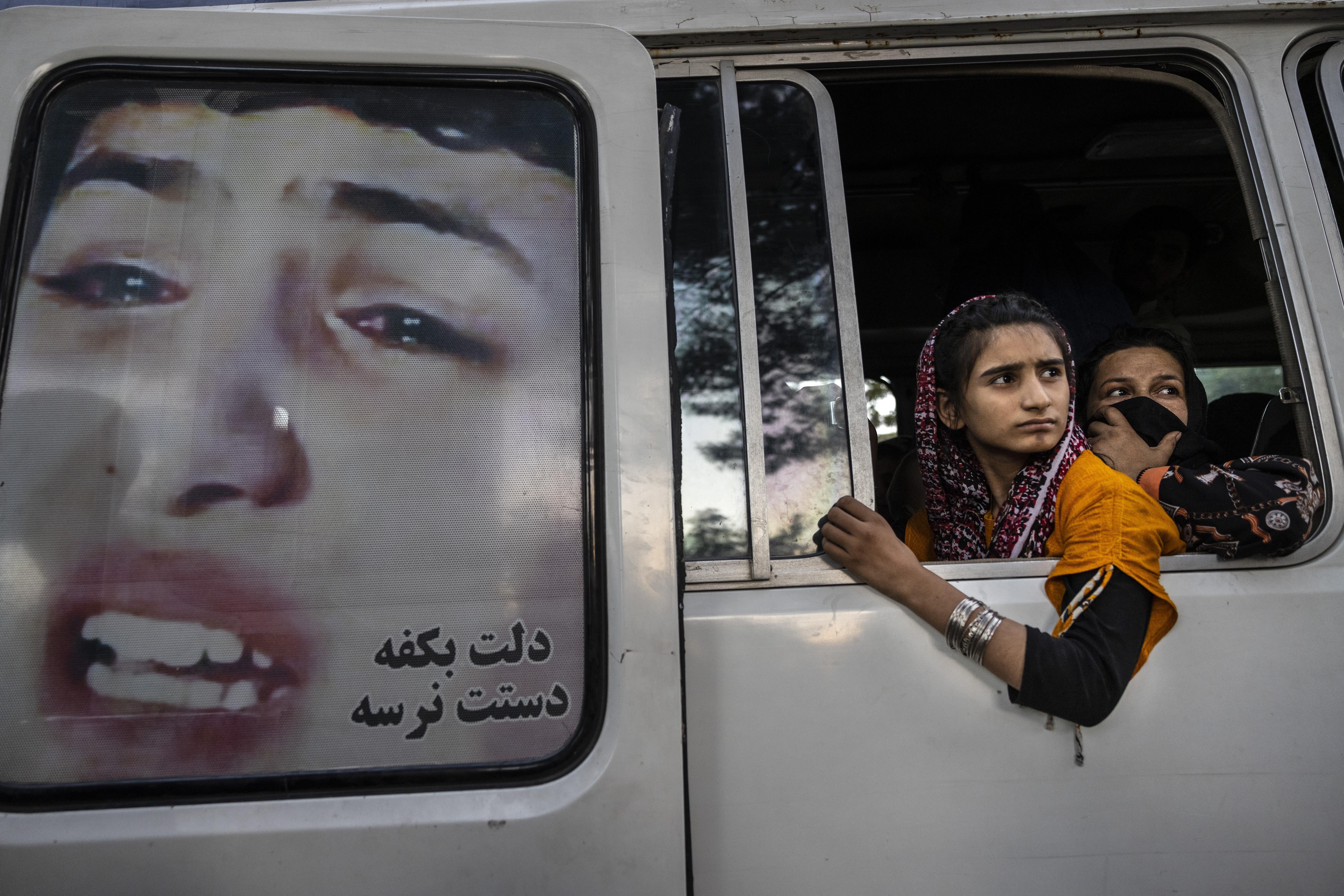 Young Afghan women look out the window of a van. There is an image of a crying woman on one of the van doors.
