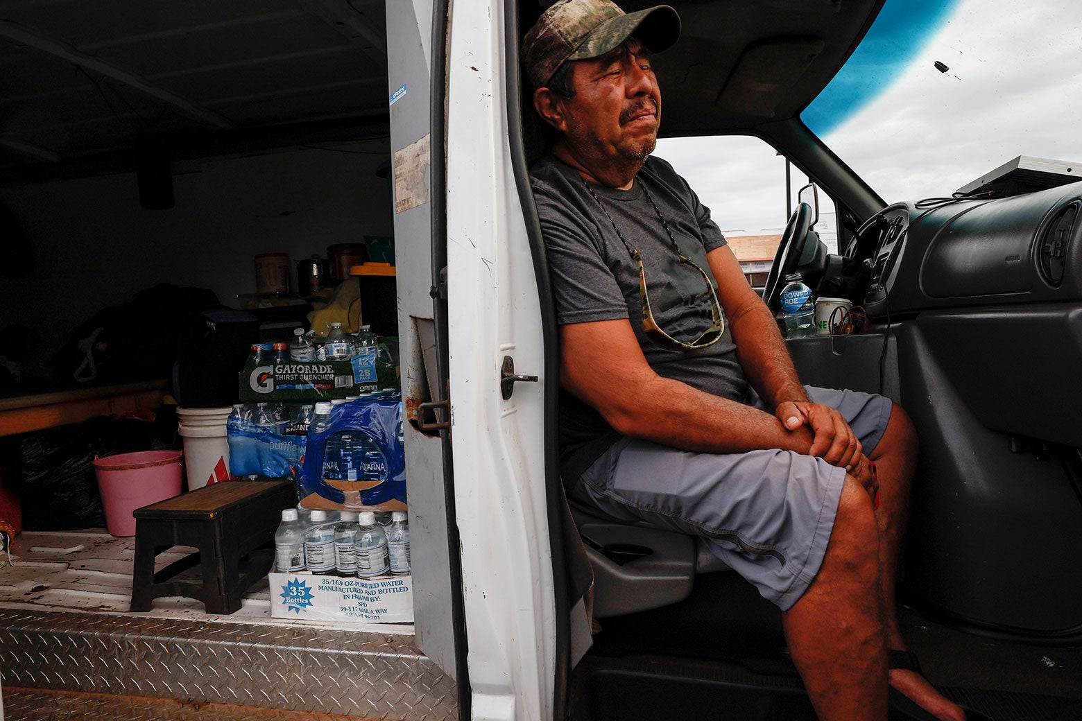 A middle-aged dark-haired man wears a green camouflage hat, T-shirt, and shorts and sits inside the cab of a truck. He looks pained and closes his eyes to stop from crying.