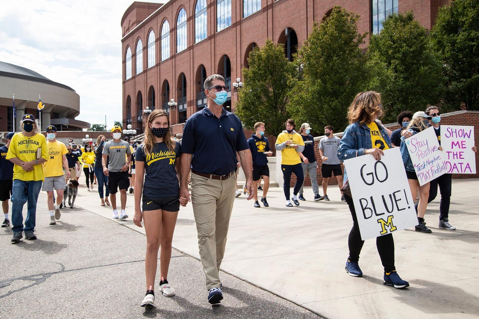 Harbaugh and a small crowd of individuals, all wearing protective masks and holding signs, walk in front of the University of Michigan's football stadium.
