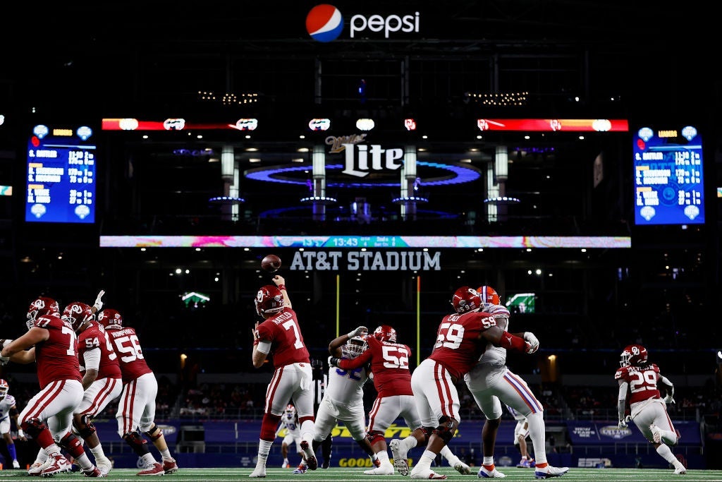 Rattler throwing a pass amid a number of offensive and defensive linemen, with logos for Goodyear, AT&T, Miller Lite, and Pepsi gleaming above against the dark backdrop of the stadium.