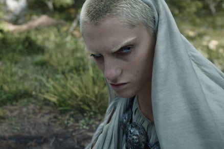 A white-cloaked figure with short-cropped, bleach-blonde hair and eyebrows scowls.