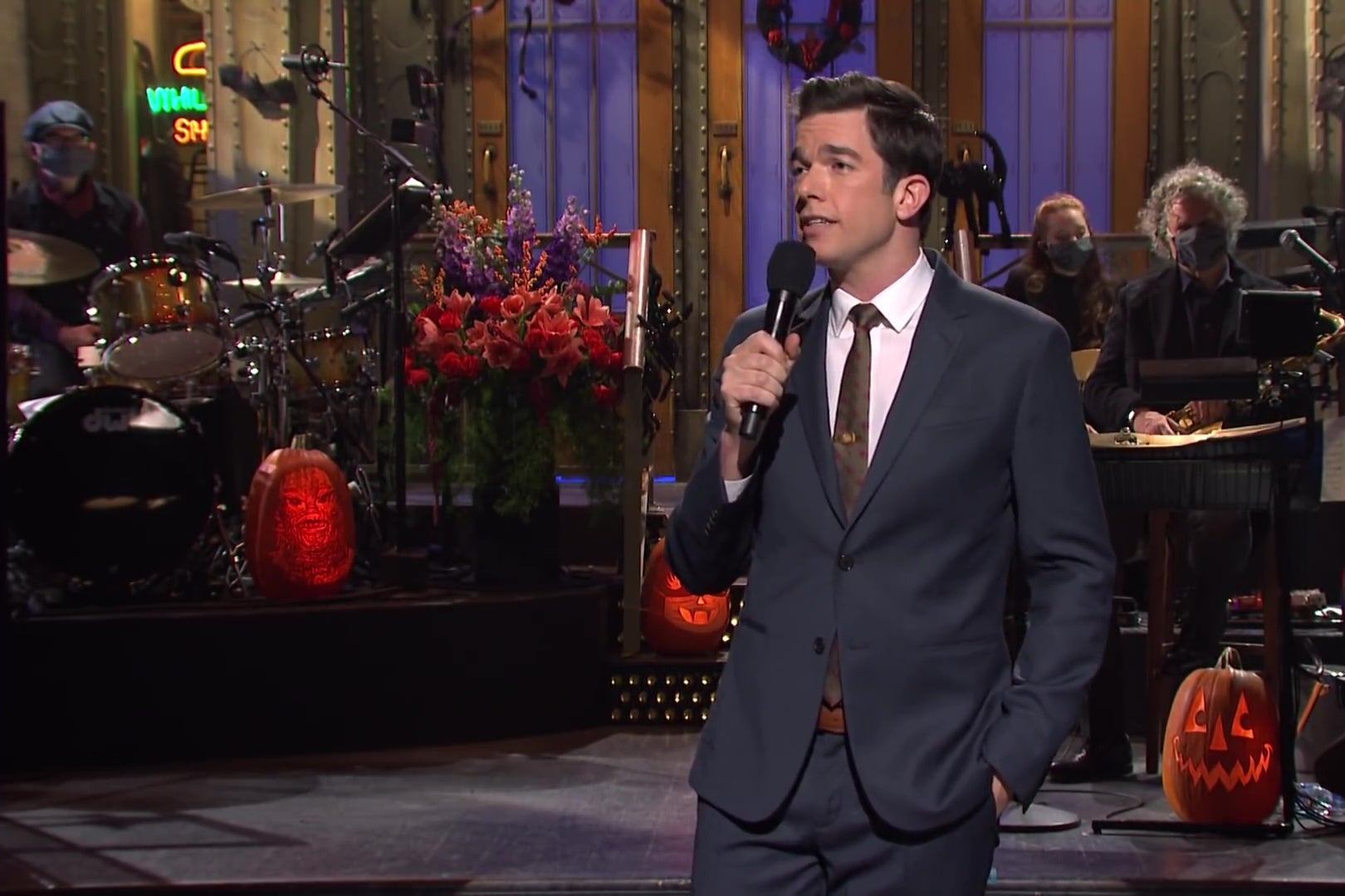 John Mulaney standing on the SNL main stage, in a grey suit, holding a microphone and telling a joke.
