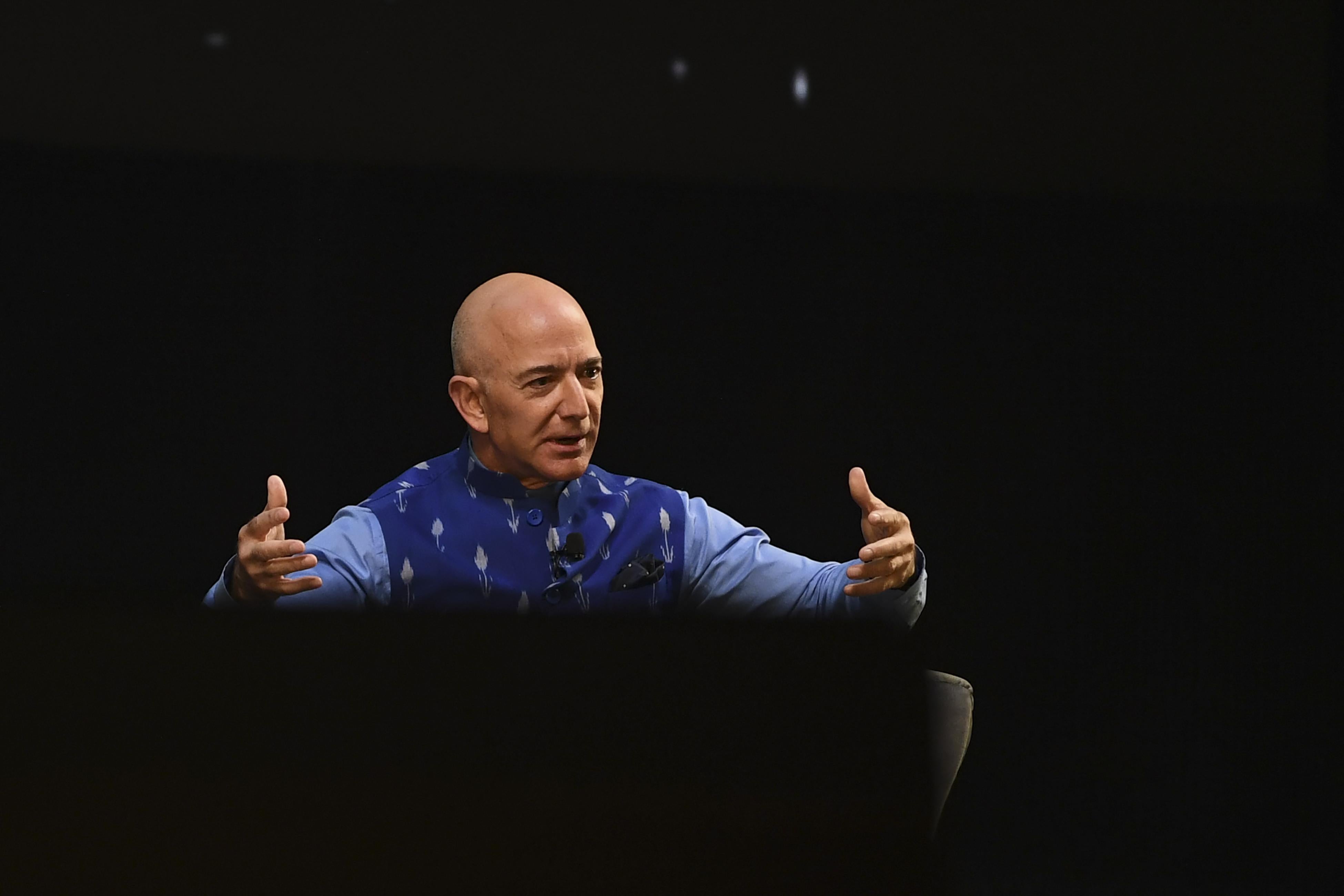 Jeff Bezos gestures with open arms as he speaks on a black stage.