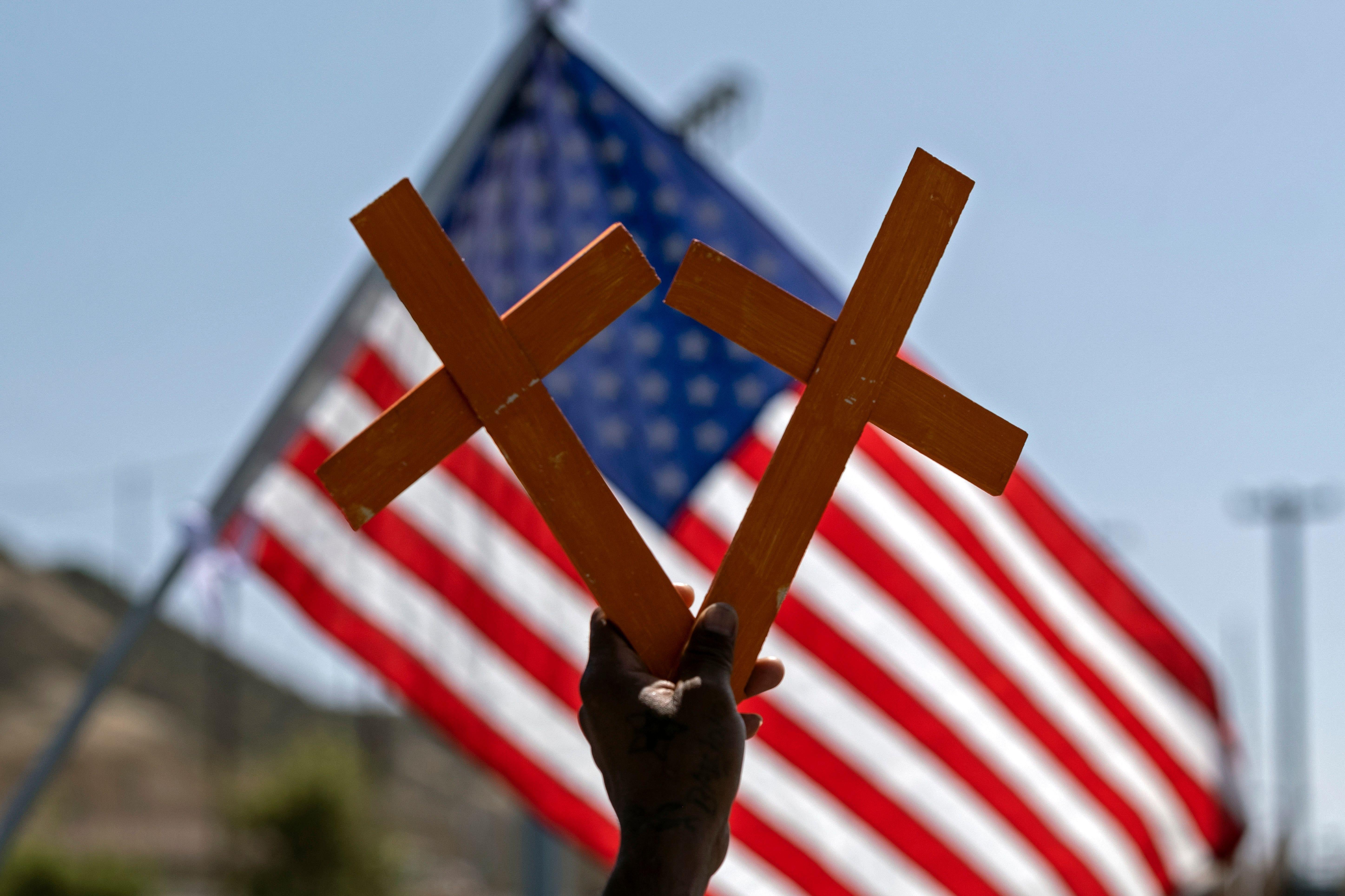 A hand holds two crosses in front of a U.S. flag.