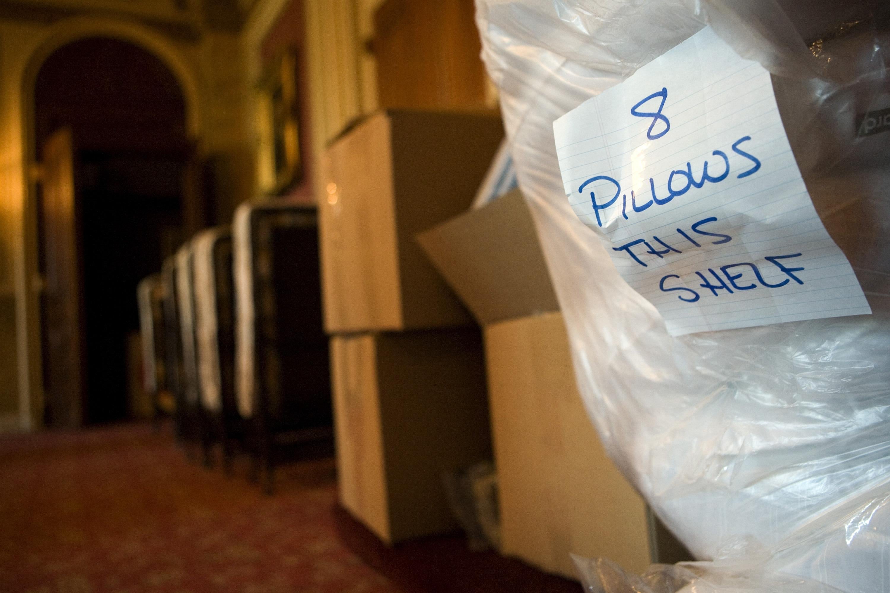 A piece of lined paper that says "8 pillows this shelf" taped to a large plastic bag of bedding next to some cardboard boxes in a hallway