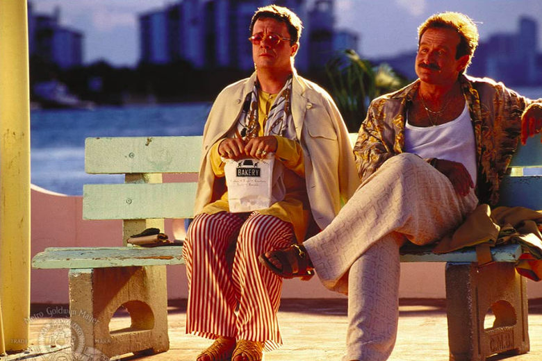 Nathan Lane, clutching a bag labeled "Bakery," sits on a park bench next to a mustached Robin Williams.