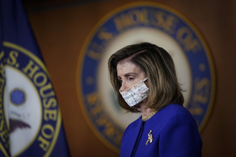 Nancy Pelosi, wearing a face mask, stands next to a House of Representatives insignia and flag.