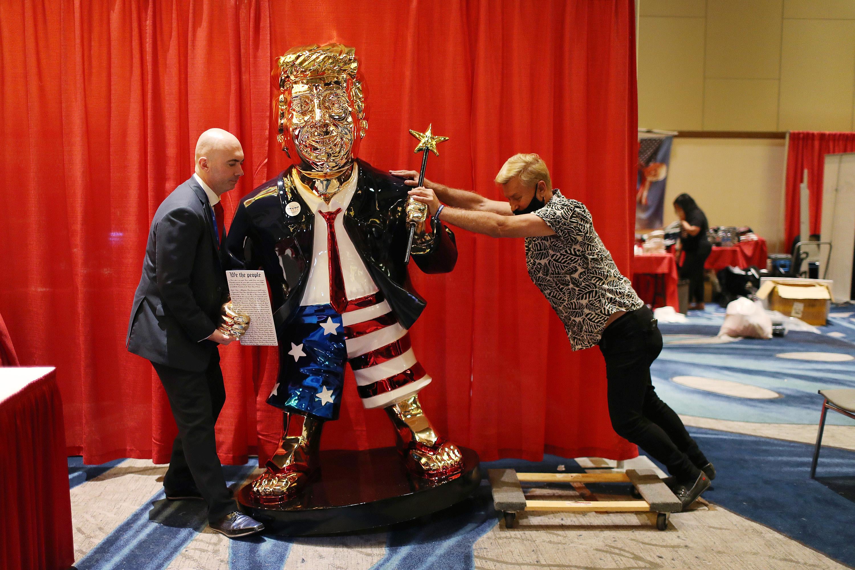 One man pushes one side of the golden Trump statue while the other man pulls from the other side. The statue depicts Trump wearing American flag shorts and holding a magic wand.